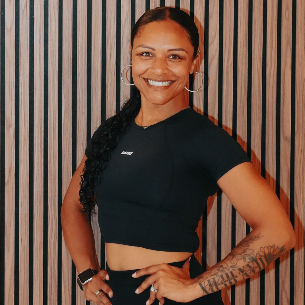 A smiling woman with long dark curly hair, wearing hoop earrings, a black cropped t-shirt, and sports leggings, standing in front of a wooden slat wall, with tattoos on her left arm.