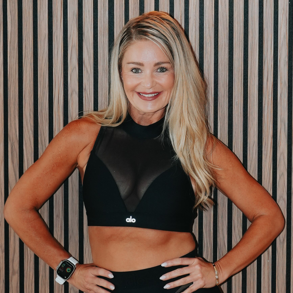 A woman with long blonde hair and a bright smile posing indoors against a wood-paneled wall, wearing a black sleeveless athletic crop top and a fitness tracker watch.