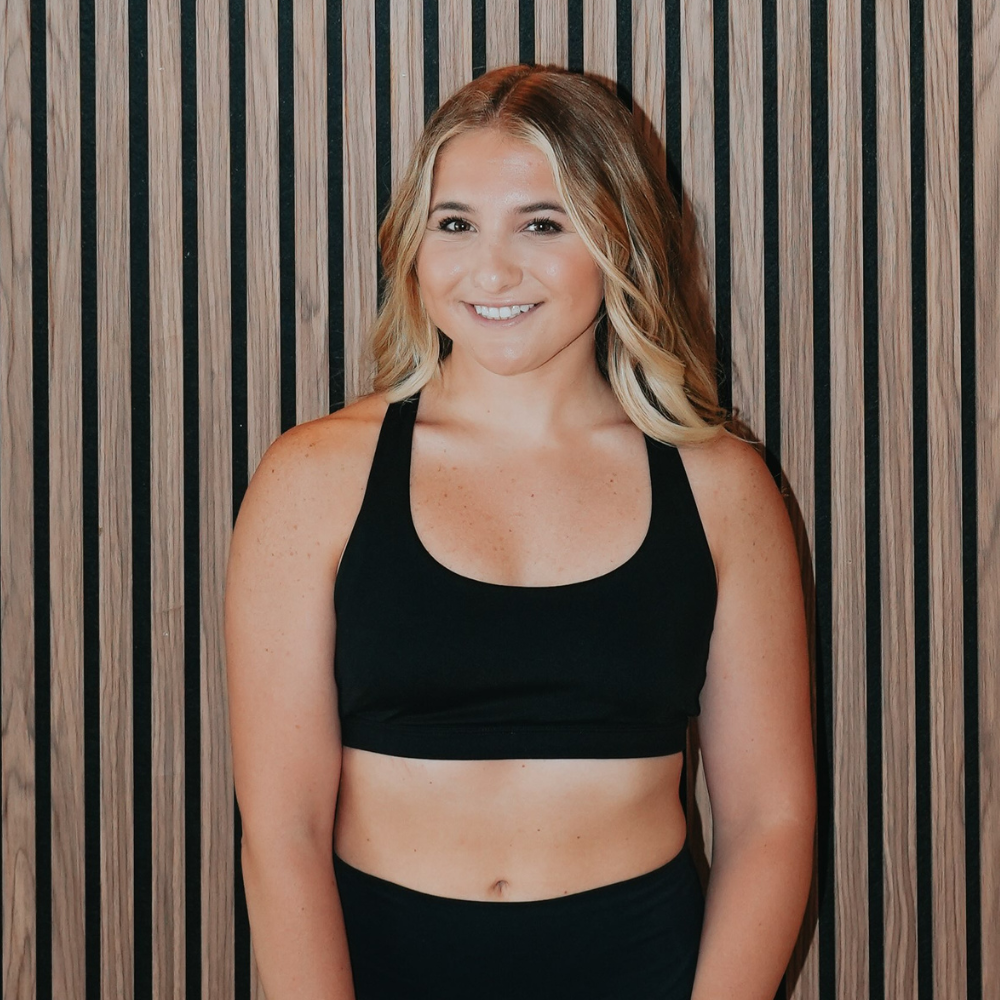 Young woman with blonde hair in loose waves, wearing a black sports bra, smiling, standing in front of a wooden slat wall.
