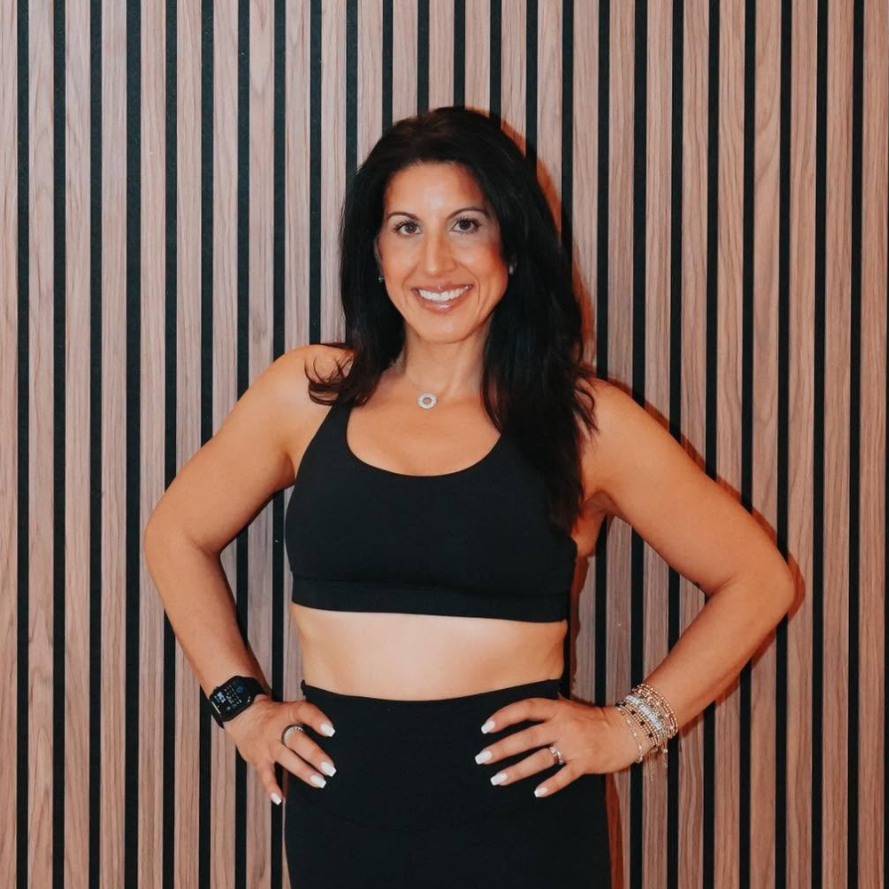 Woman with black hair wearing a black sports bra and matching high-waisted leggings, smiling and standing confidently with her hands on her hips against a wooden slat background.