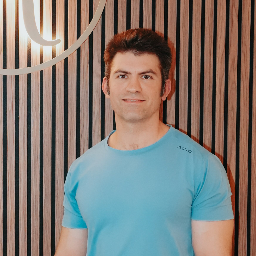 A man with dark hair, wearing a light blue T-shirt, standing in front of a wooden wall with vertical slats and a wall lamp to the left.