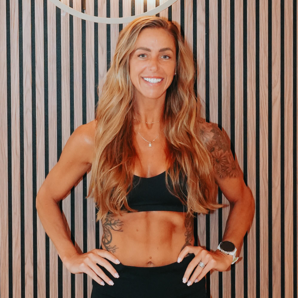 A fit woman with long, wavy blonde hair, wearing a black sports bra, tattoos on her arms and side, smiling, standing with hands on hips in front of a striped wooden wall.