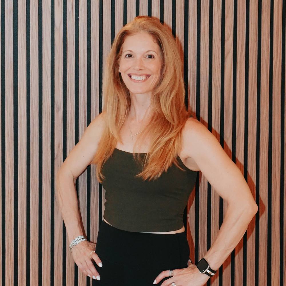 A woman with long red hair smiling, wearing a black sleeveless top and black pants, standing with her hands on her hips in front of a wooden slat wall.