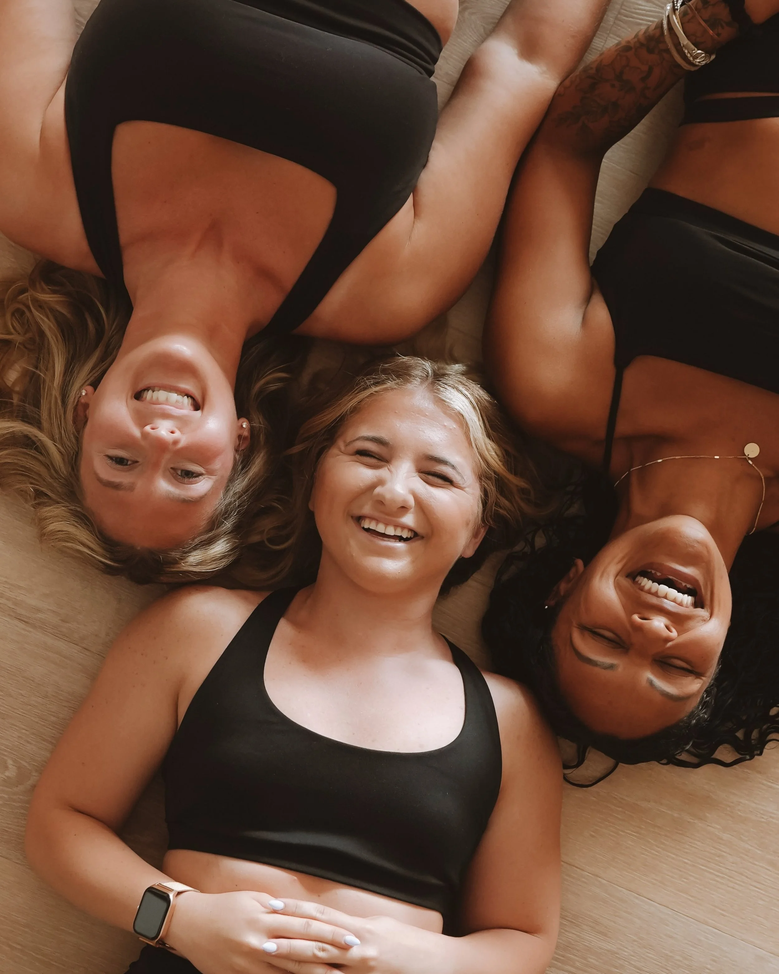 Four women lying on the floor close together, smiling and looking up at the camera, with their heads close together and their bodies forming an abstract pattern.