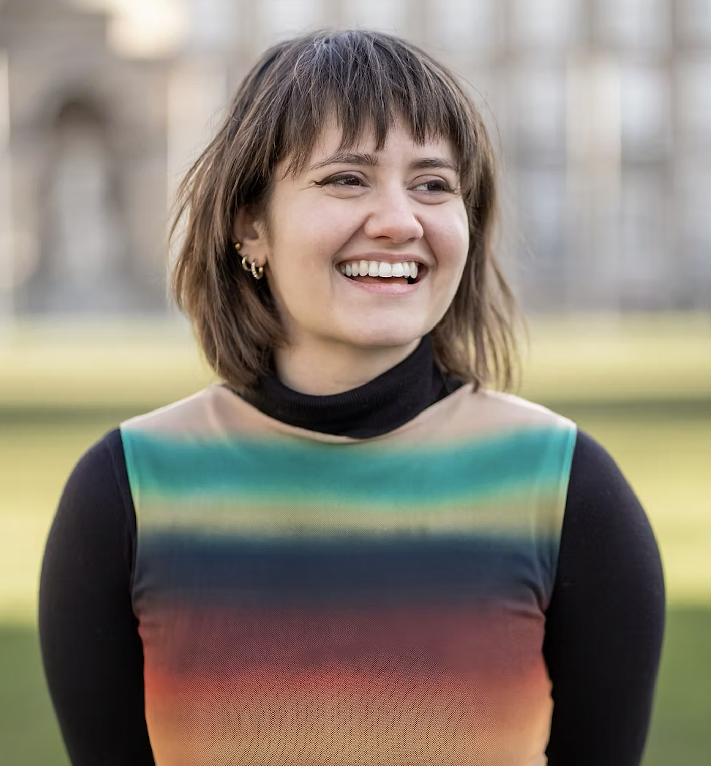 A woman with short brown hair and multiple earrings smiling outdoors, wearing a colorful gradient top and black turtleneck.