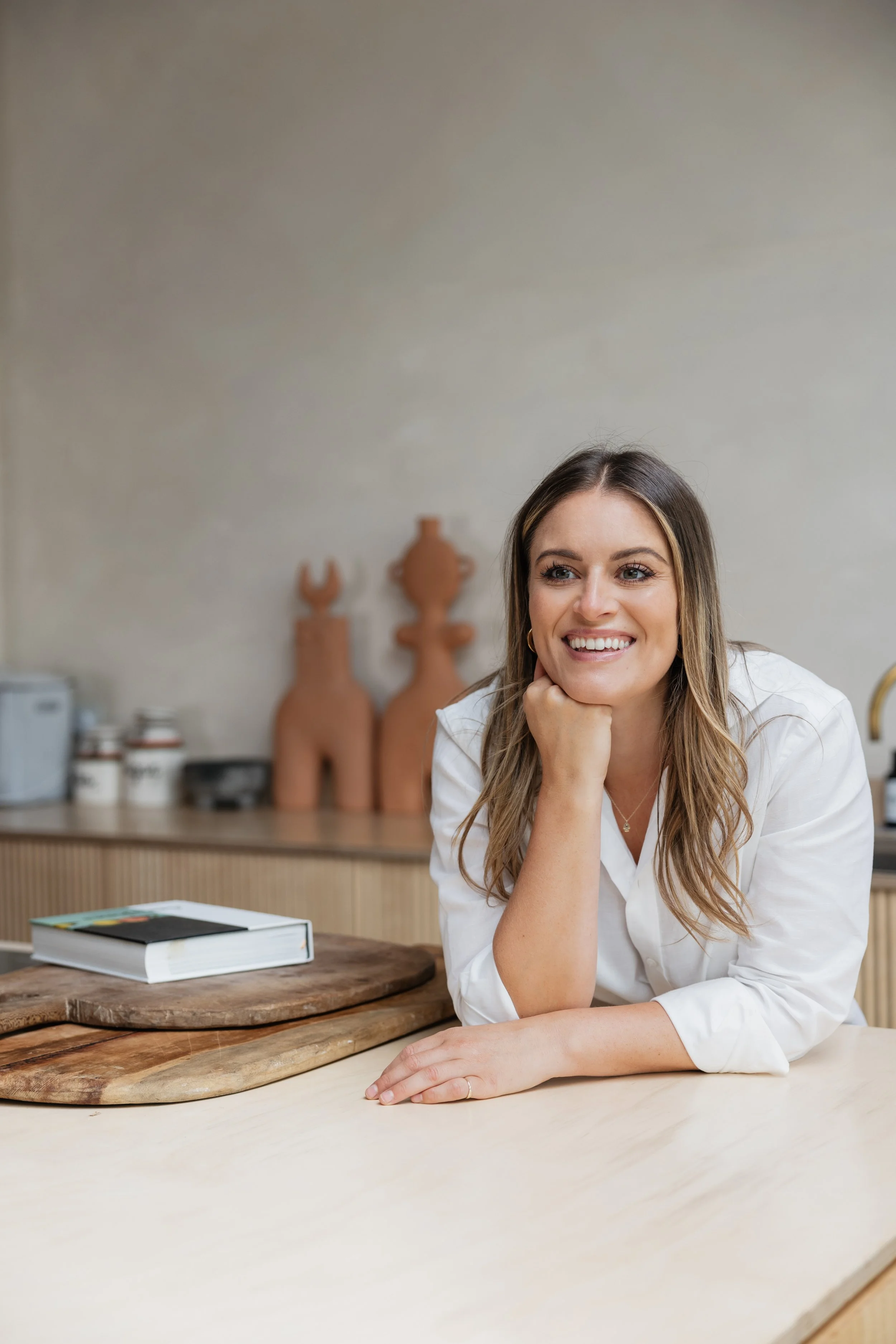 A woman smiling and resting her head on her hand at a kitchen counter, with a book and a wooden cutting board in front of her.