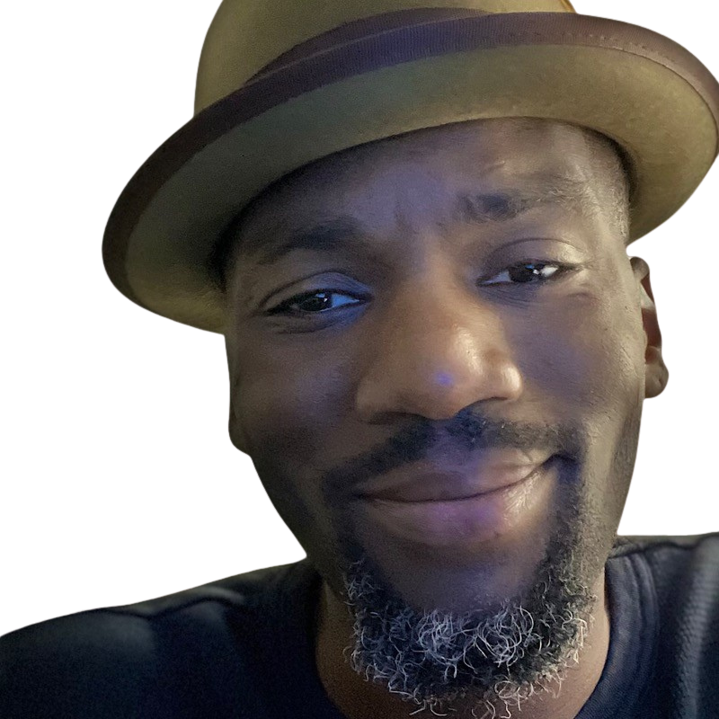 Close-up of a smiling man wearing a tan fedora hat and a black shirt, with a visible beard and mustache.