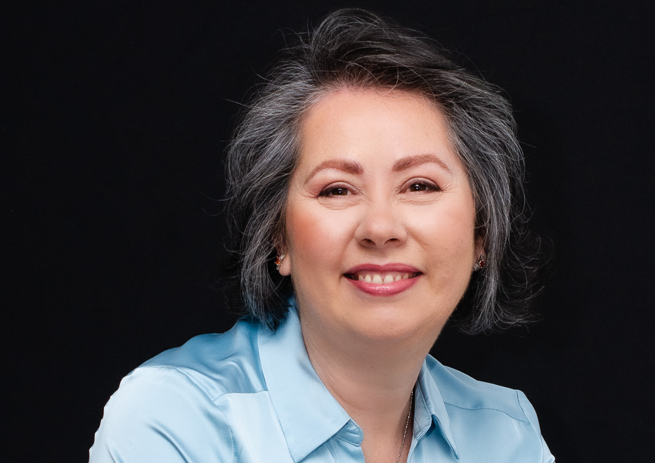 A smiling middle-aged woman with gray hair wearing a light blue blouse against a black background.