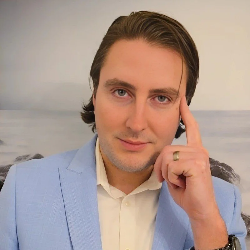 A man with dark hair styled to the side, wearing a light blue blazer and white shirt, with his right hand touching his temple, standing in front of a background with the ocean and rocks.