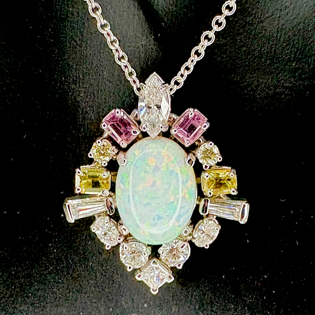 A white gold large oval opal centerpiece surrounded by pink, yellow, and clear gemstones hand fabricated pendant on a white gold chain.