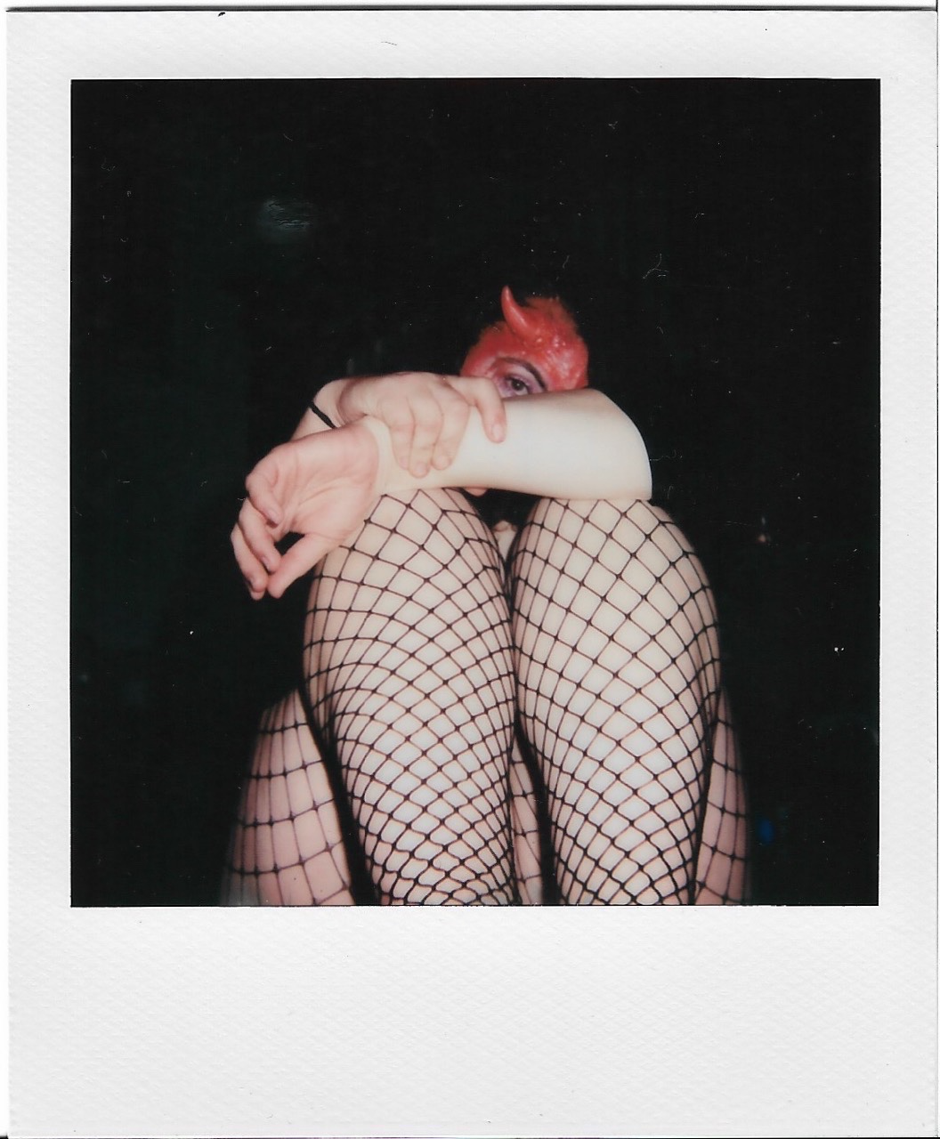 A person with red horns and face paint, wearing fishnet stockings, sitting with knees drawn up and arms wrapped around their legs, against a dark background.