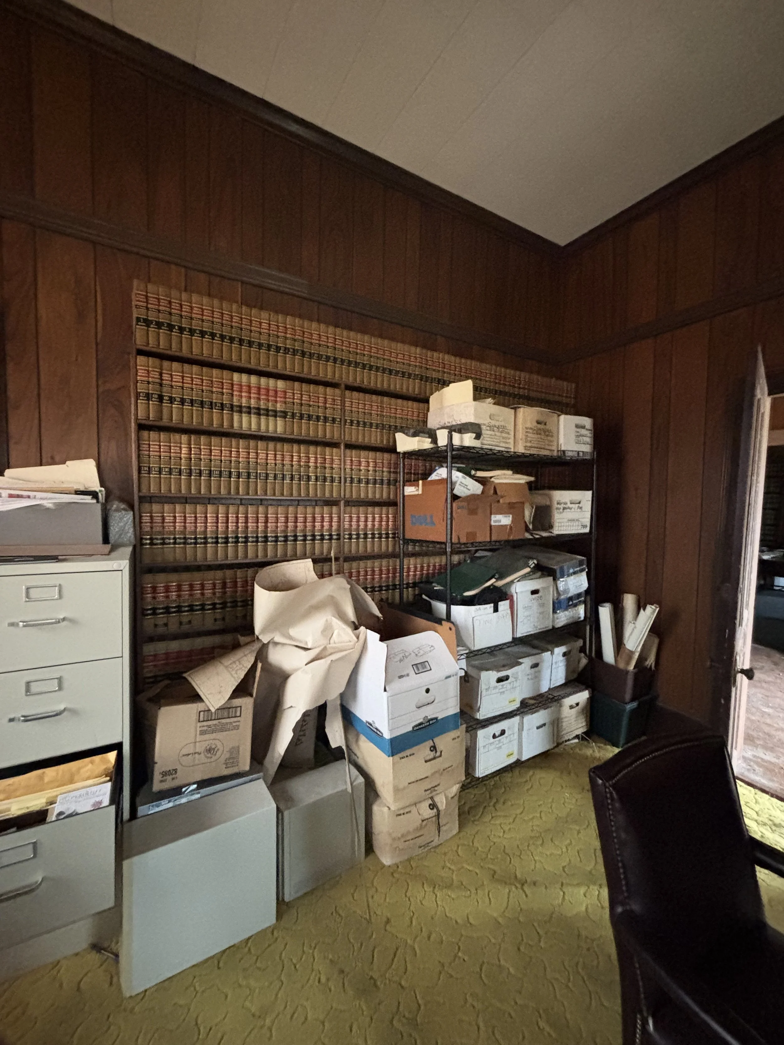 Office with wood-paneled walls, a metal shelving unit filled with boxes and files, a beige filing cabinet, and a black leather chair on yellow patterned carpet.