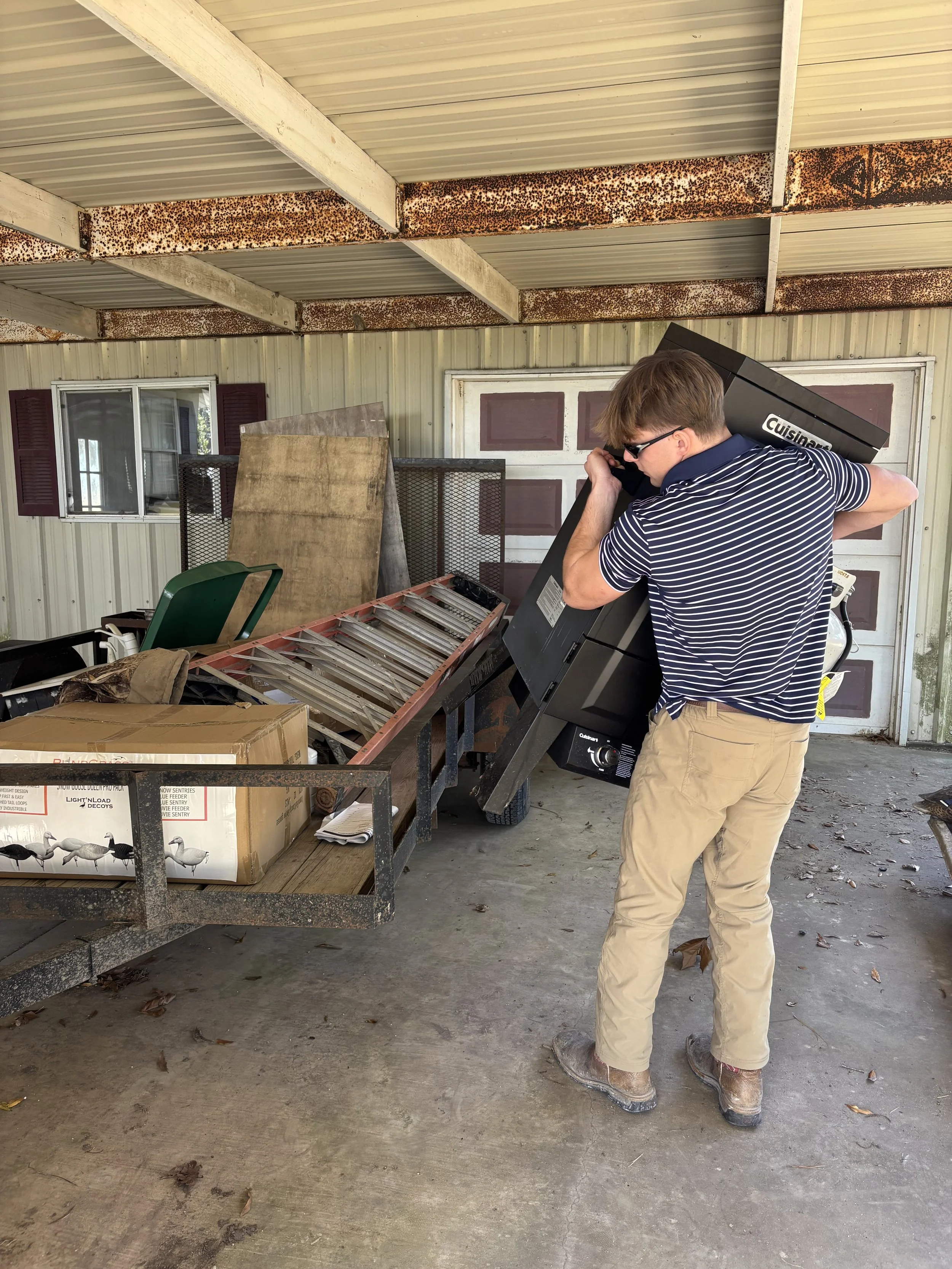A man in a striped shirt and khaki pants moving a large black  grill into or out of a trailer in a garage or shed with a rusty roof and a window.