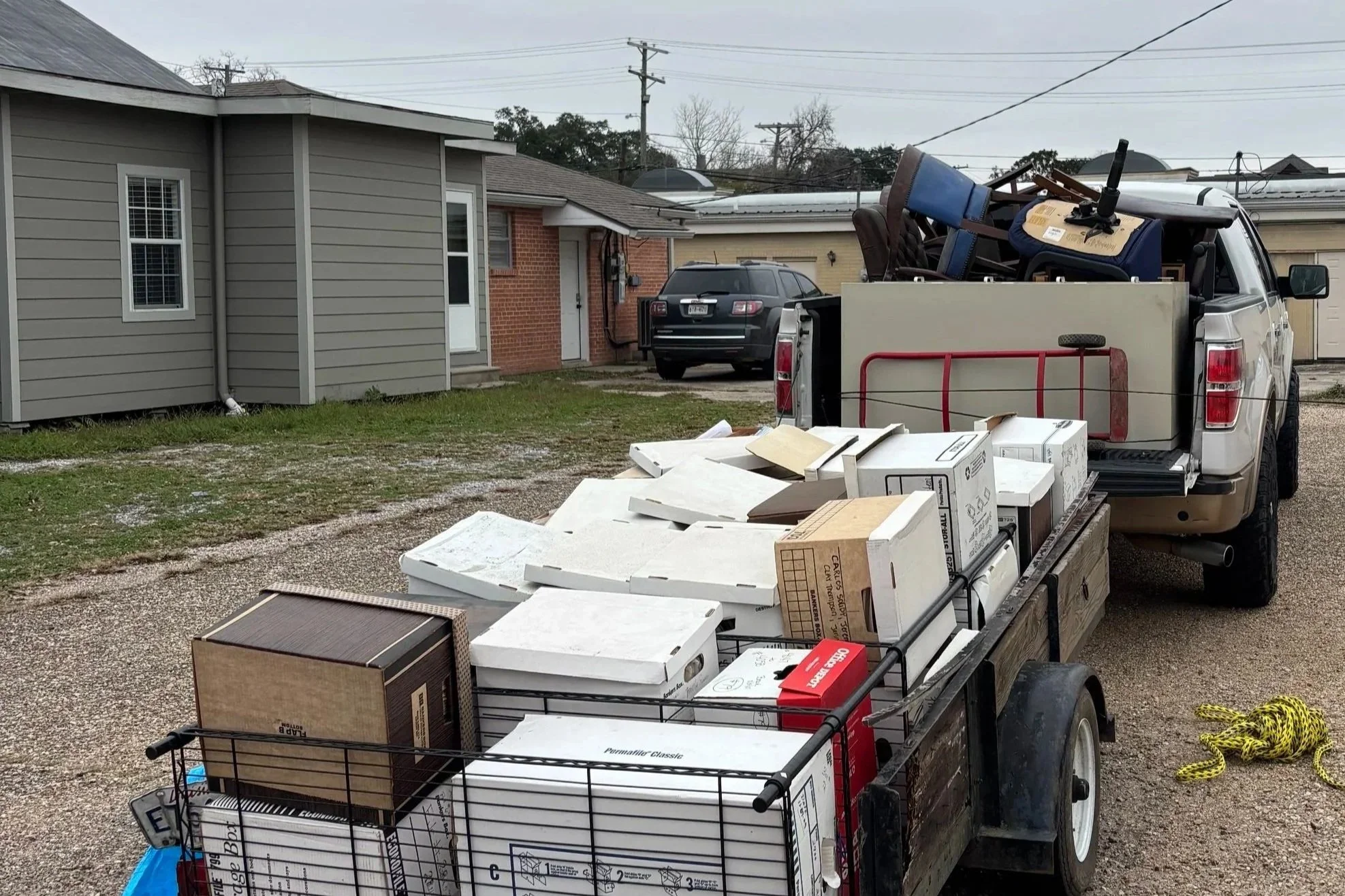 A pickup truck with its bed loaded with various furniture, boxes, and household items parked in a driveway in front of residential houses.