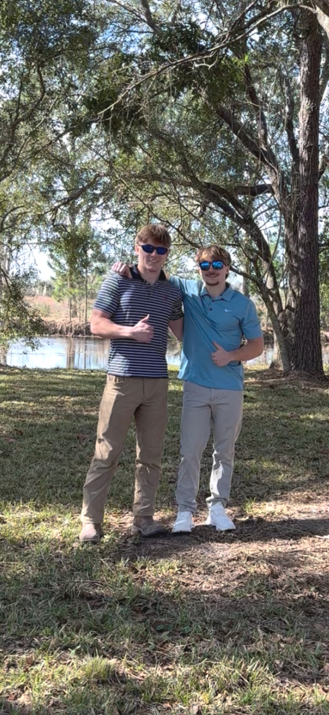 Two young men standing outdoors under a large tree on grass, smiling and giving thumbs-up gestures. One is wearing a striped polo shirt and beige pants, the other is wearing a light blue shirt and khaki pants. They are near a body of water and the background shows more trees.