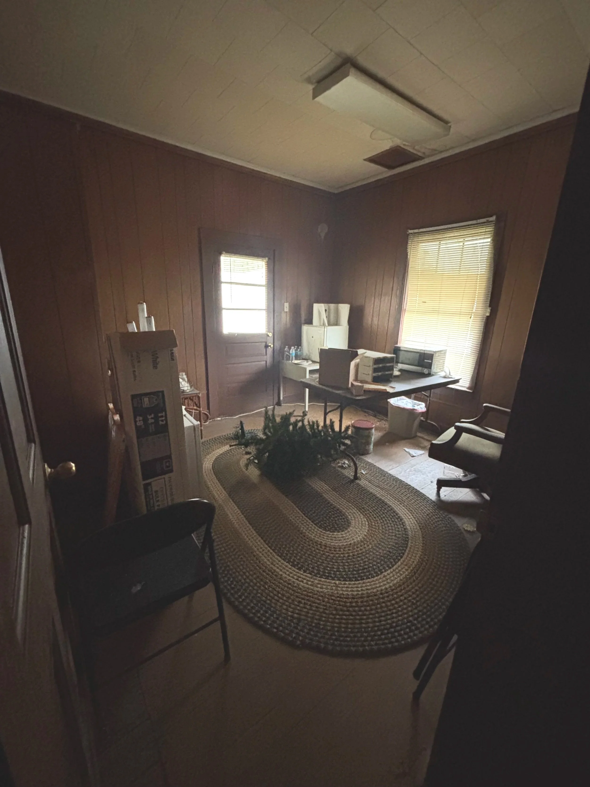 A small, cluttered office with wood-paneled walls, a window with blinds, and a door. There is a dark rug on the floor, topped with pulled-down Christmas tree. A desk with office supplies, boxes, and a microwave is near the window. Two chairs are positioned in the room, and there are some boxes and miscellaneous items around.