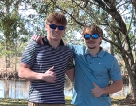 Two young men standing outdoors near a lake, smiling and giving thumbs-up hand signals, with trees in the background.