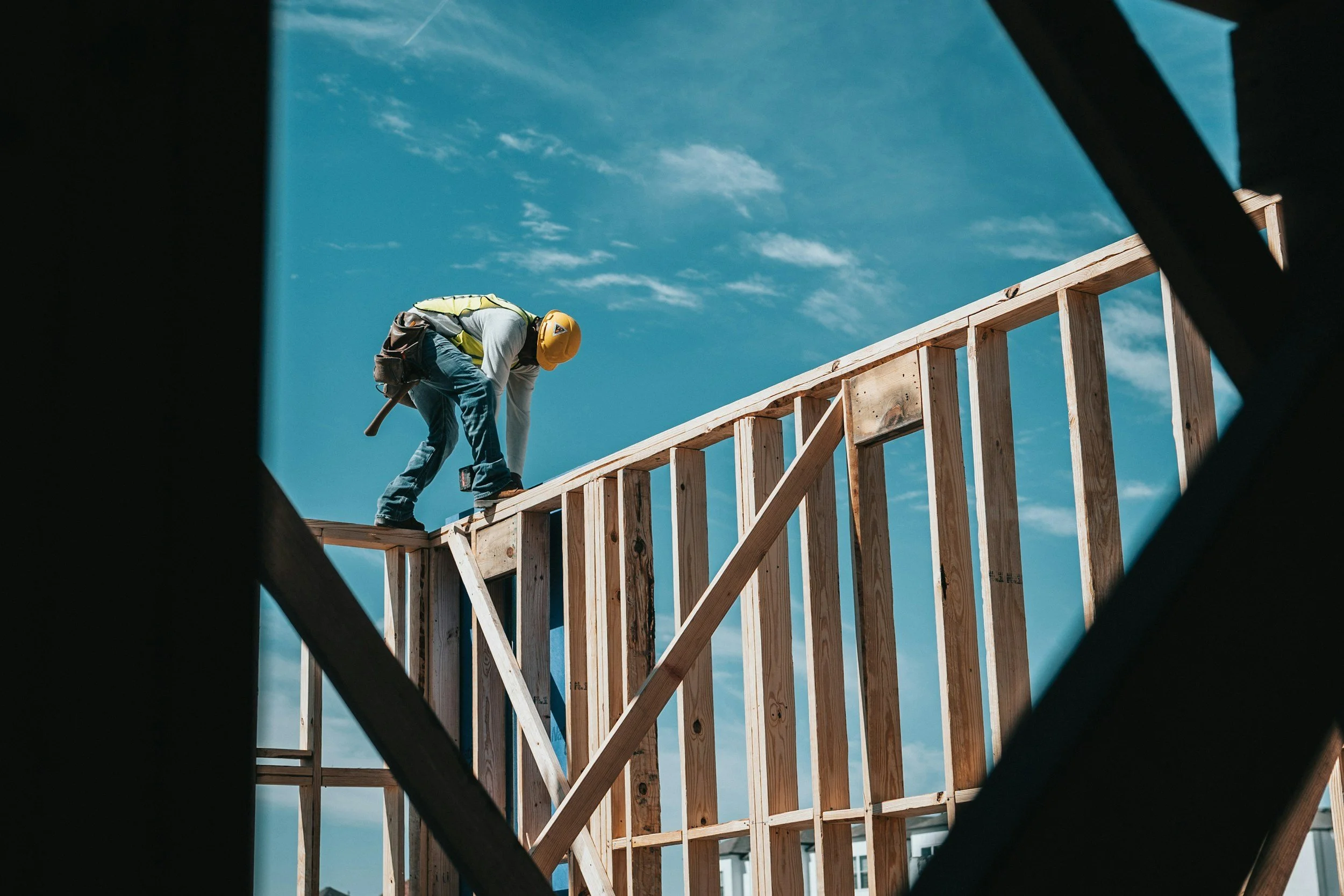 A construction worker wearing a yellow hard hat and a safety vest, bending over and working on a wooden framing structure at a building site under a blue sky.