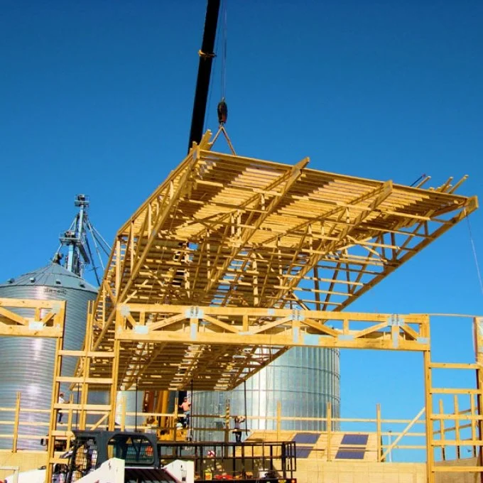 Construction site with a wooden platform and silos, crane lifting the platform into place, under clear blue sky.