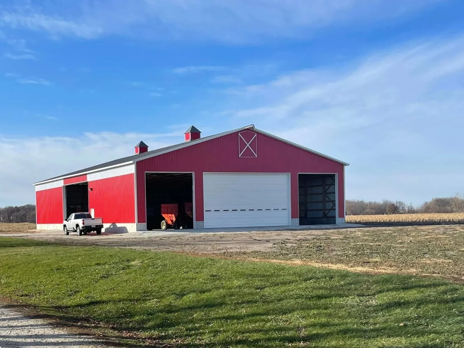 A large red barn with white trim, two small cupolas on the roof, and a white garage door, situated on a farm with a pickup truck parked nearby and open barn doors revealing farming equipment inside, under a partly cloudy blue sky.
