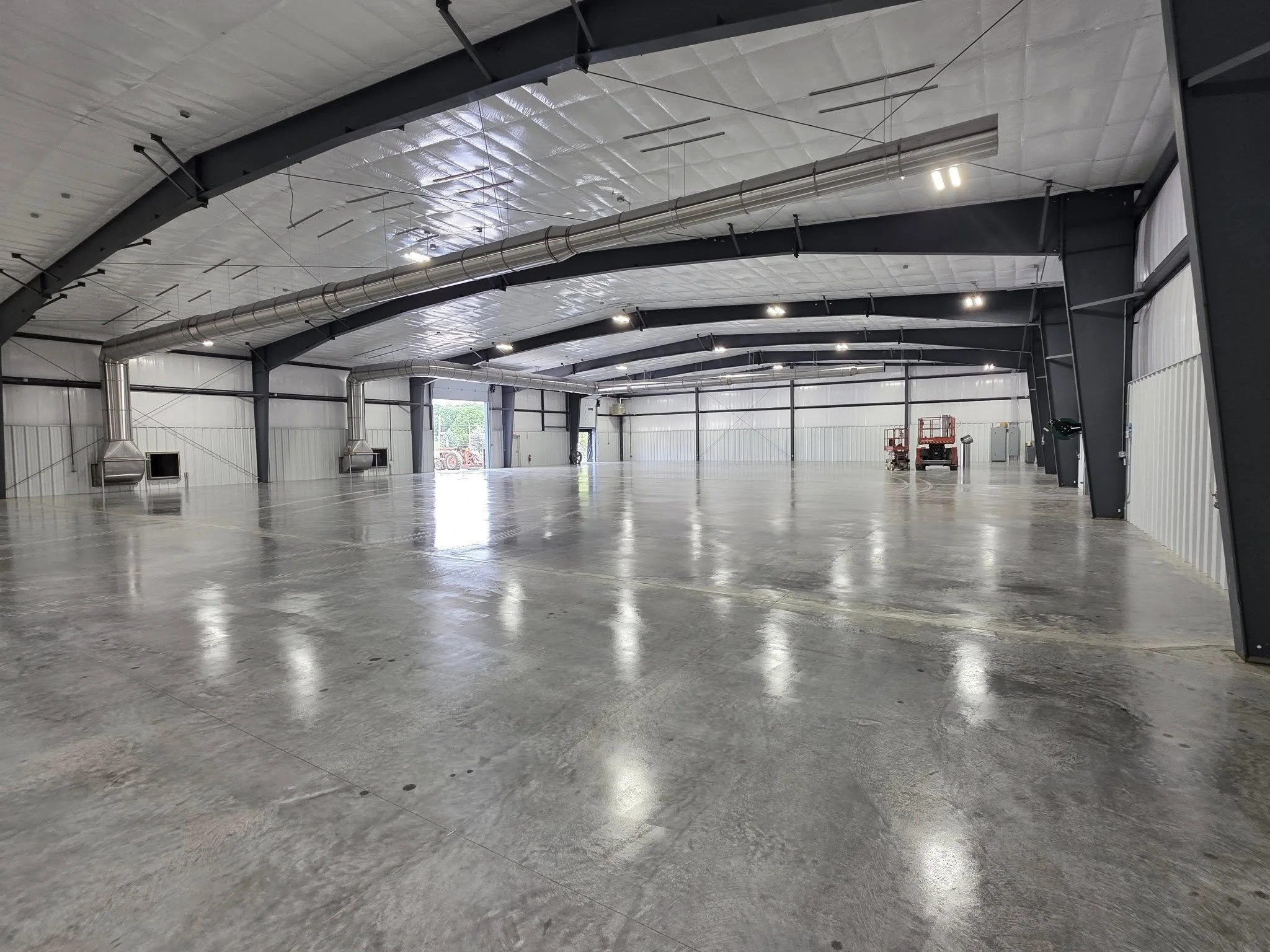 Interior of a large empty warehouse with a polished concrete floor, metal beams, and large ventilation ducts suspended from the ceiling. There are some small windows and machinery on the edges.