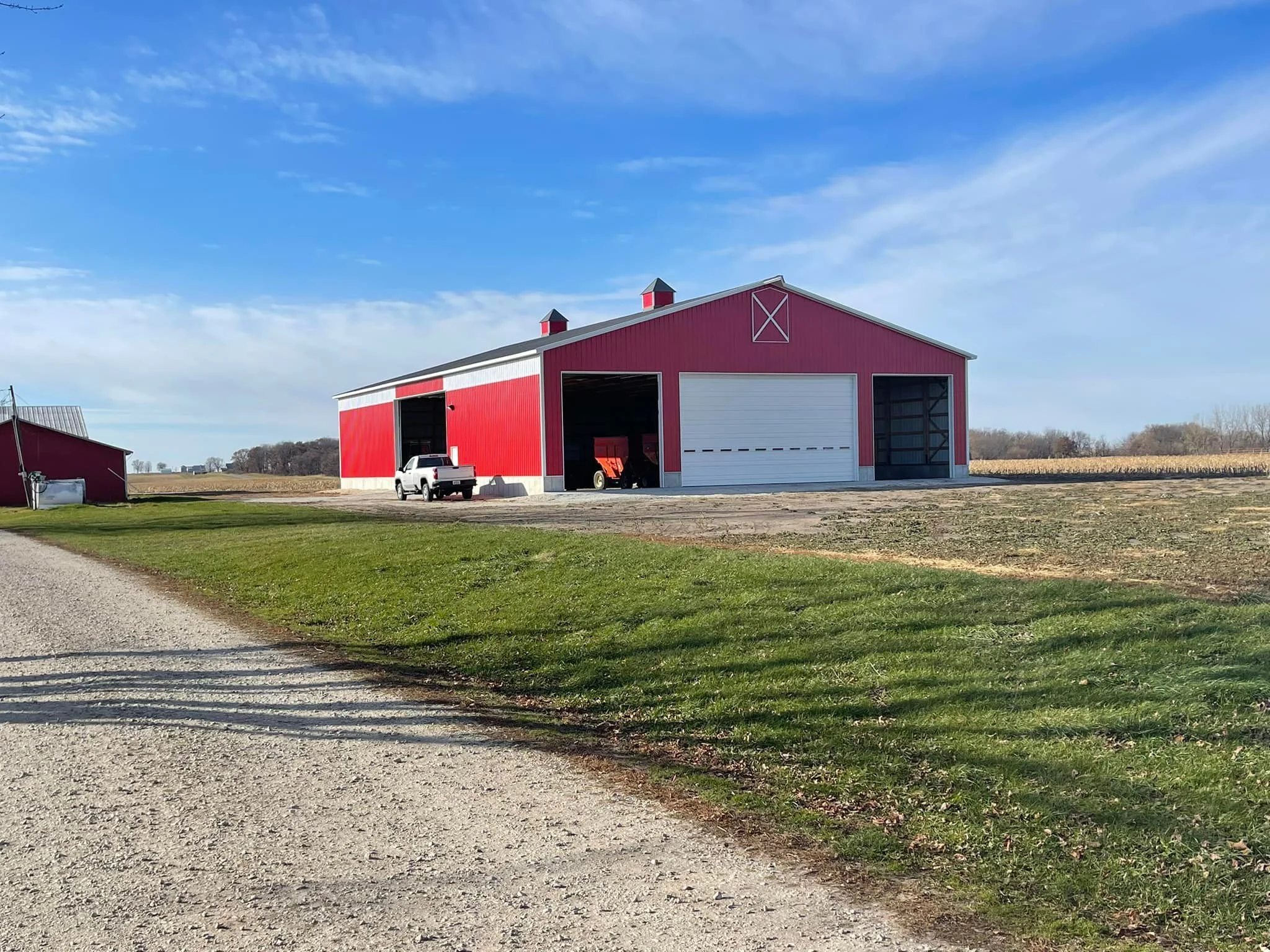 Red barn with white door and two small cupolas on the roof, parked truck and farm equipment nearby, open field and blue sky in the background.