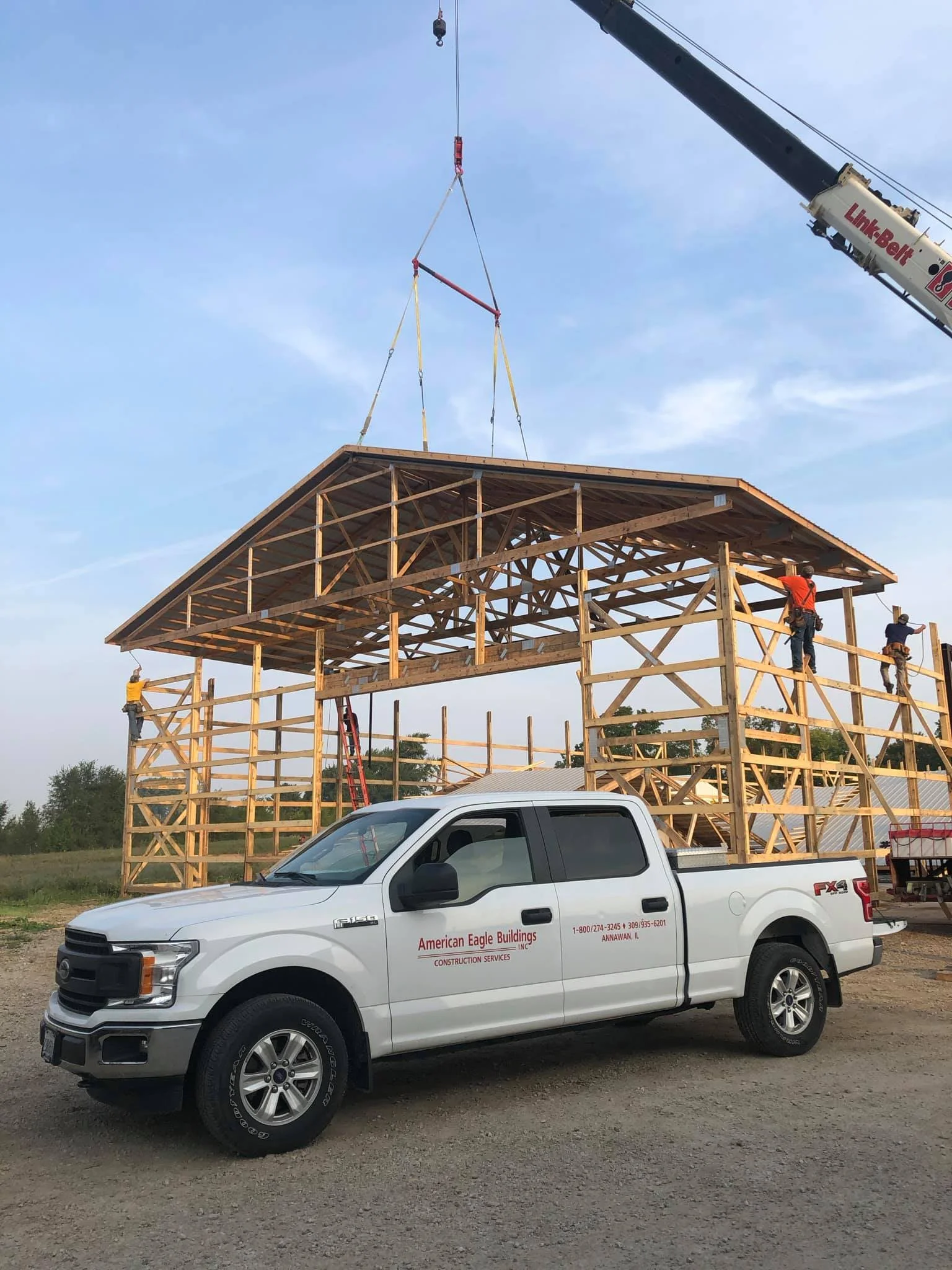 A construction site with a wooden framework structure for a building and a crane lifting materials. A white pickup truck with company information is parked in the foreground.