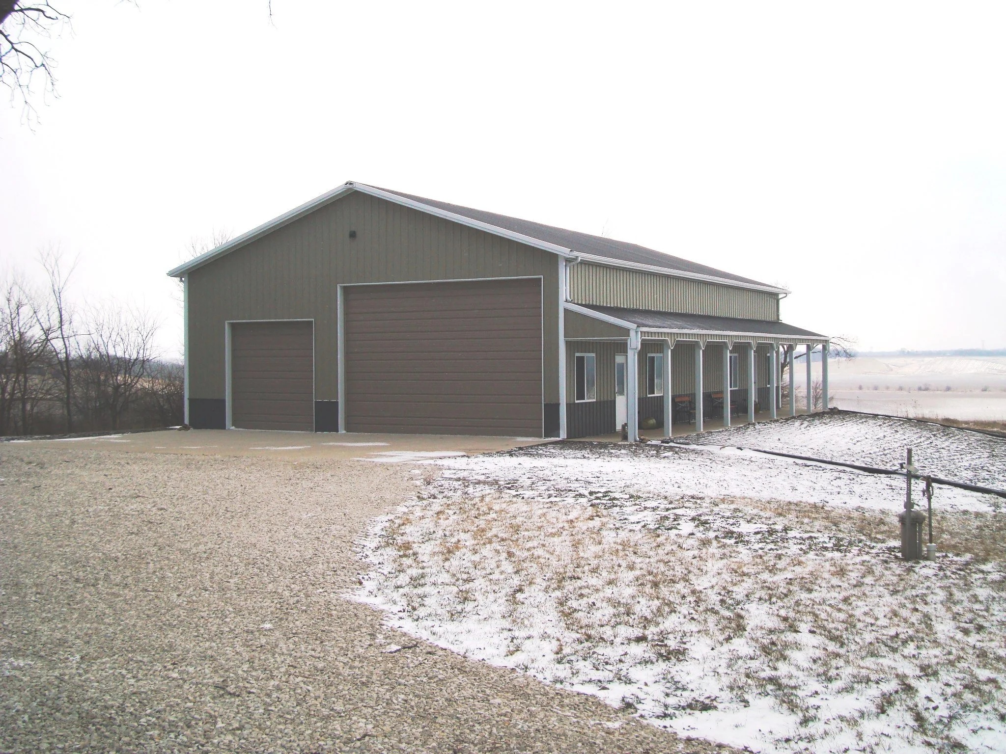 A grey barn with a gabled roof and large garage doors, set in a rural area with leafless trees and a partly snow-covered ground.