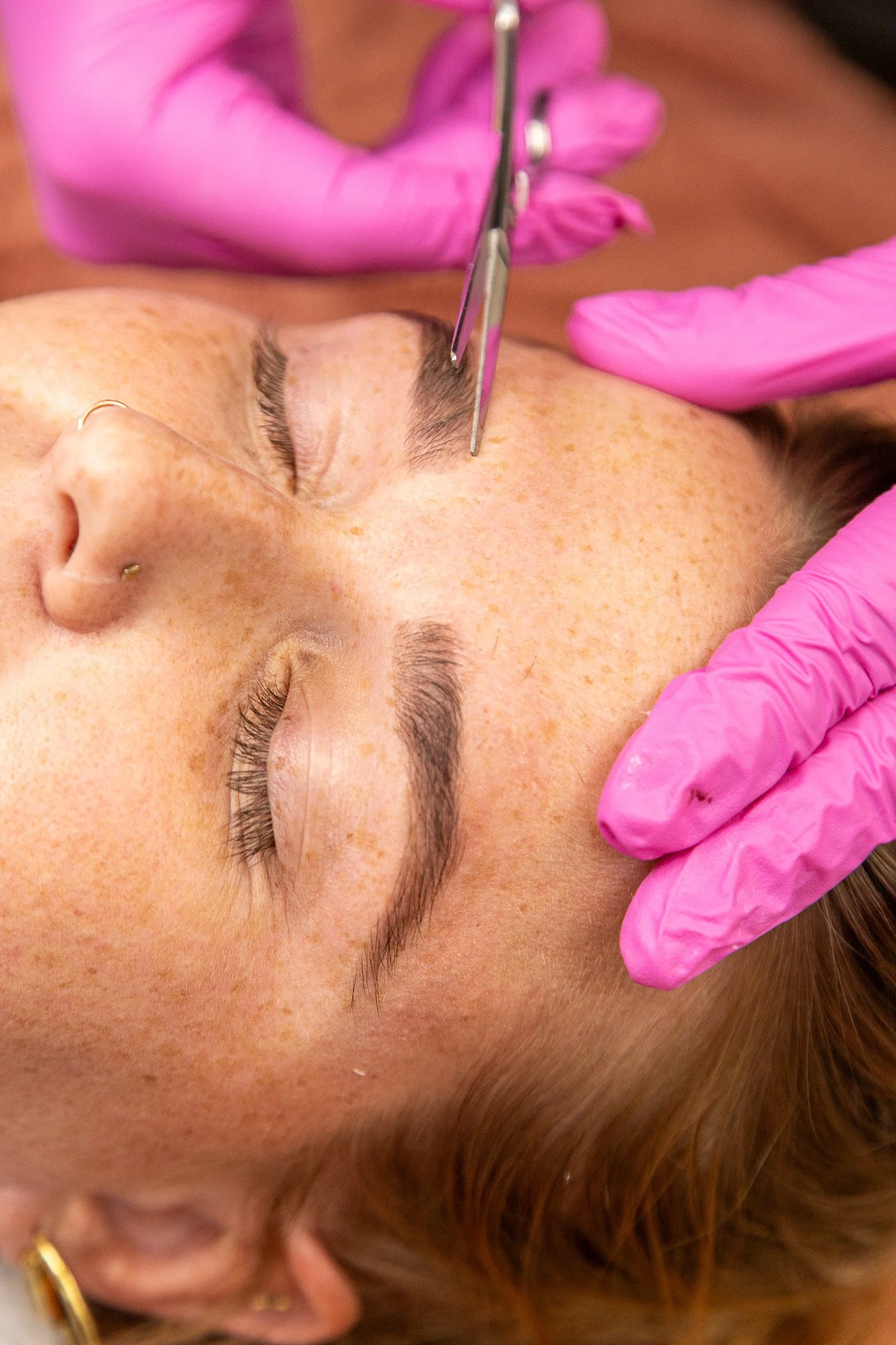 A person lying on their back during a cosmetic tattoo procedure, with closed eyes and relaxed facial expression. A practitioner wearing pink gloves is using a fine needle tool near the person's eyebrow, which appears to be tattooed or microbladed.