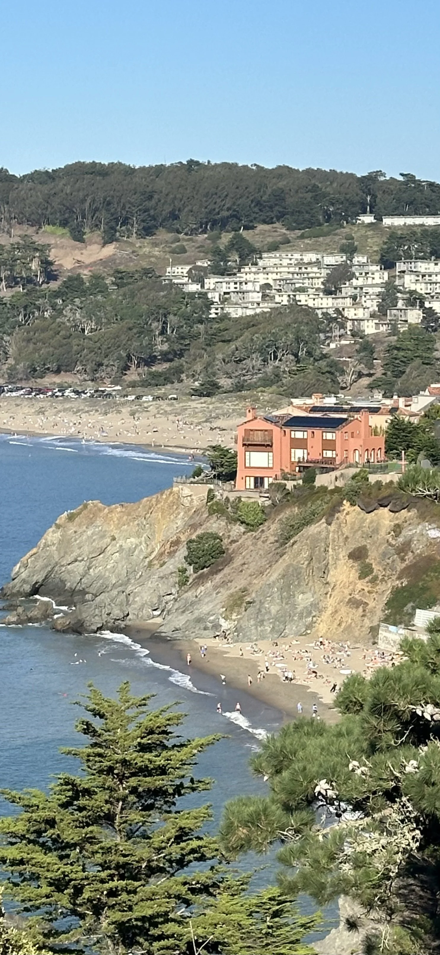A coastal scene with a beach, ocean waves, a rocky cliff with a large peach-colored house, and a hillside with numerous white residential buildings surrounded by trees.