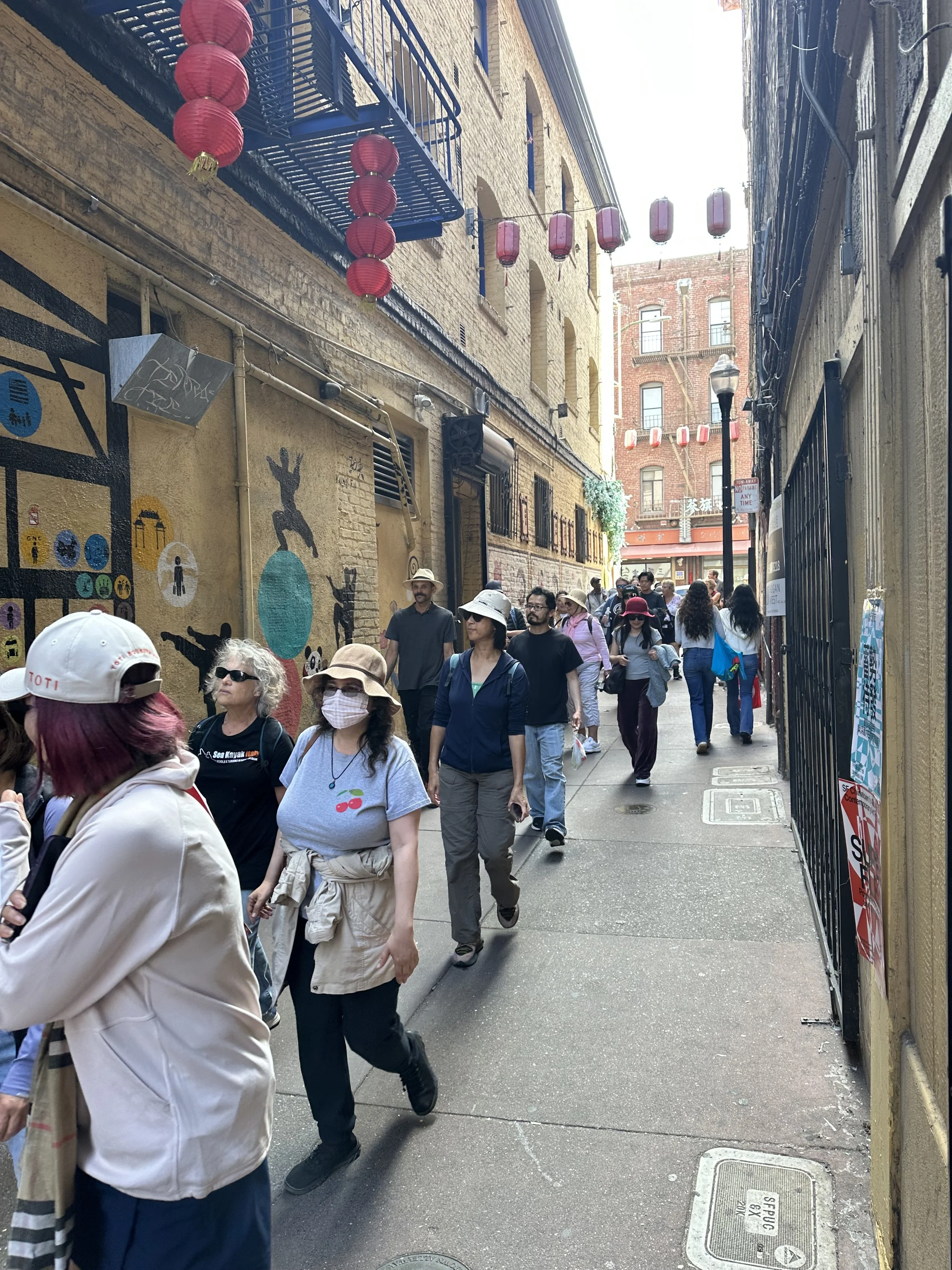 People walking through a narrow alley decorated with red lanterns, with graffiti and murals on the walls and buildings on both sides.