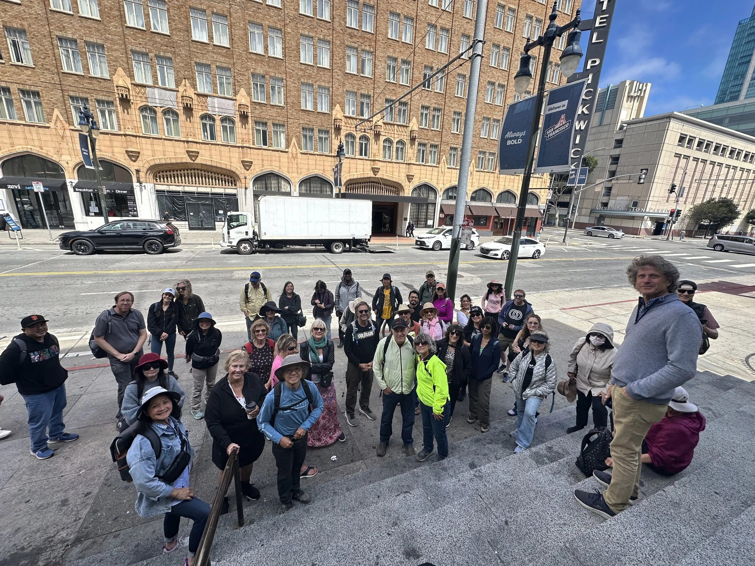 Group of tourists posing for a photo on city steps in front of a street with parked cars and tall buildings, some wearing hats, sunglasses, and backpacks.