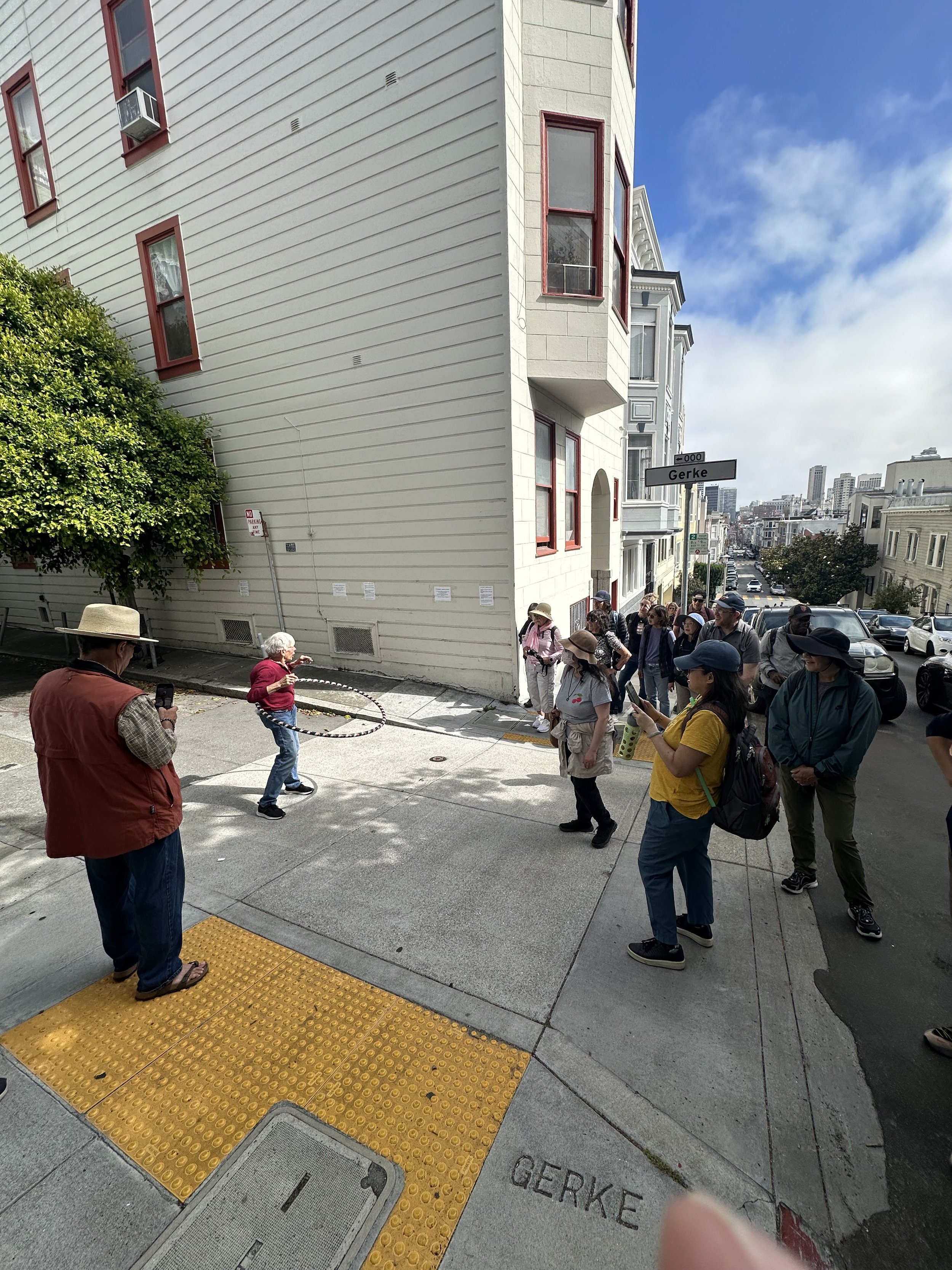 A group of people standing on a city sidewalk at the corner of Gerke street, with two adults playing a game of hula hoop and a crowd of spectators watching. Children and adults are present, some taking photos. The street is lined with buildings, and a blue sky with some clouds is visible.