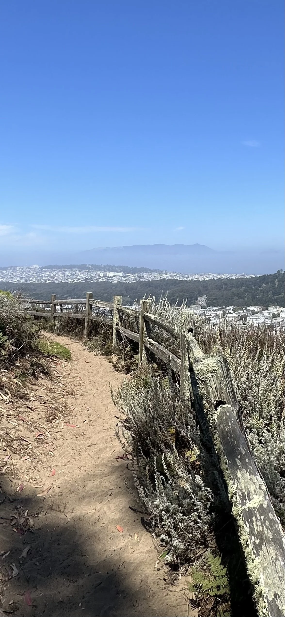 A dirt trail on a hillside with a wooden fence, leading towards a distant view of a cityscape with hills and mountains in the background under a clear blue sky.