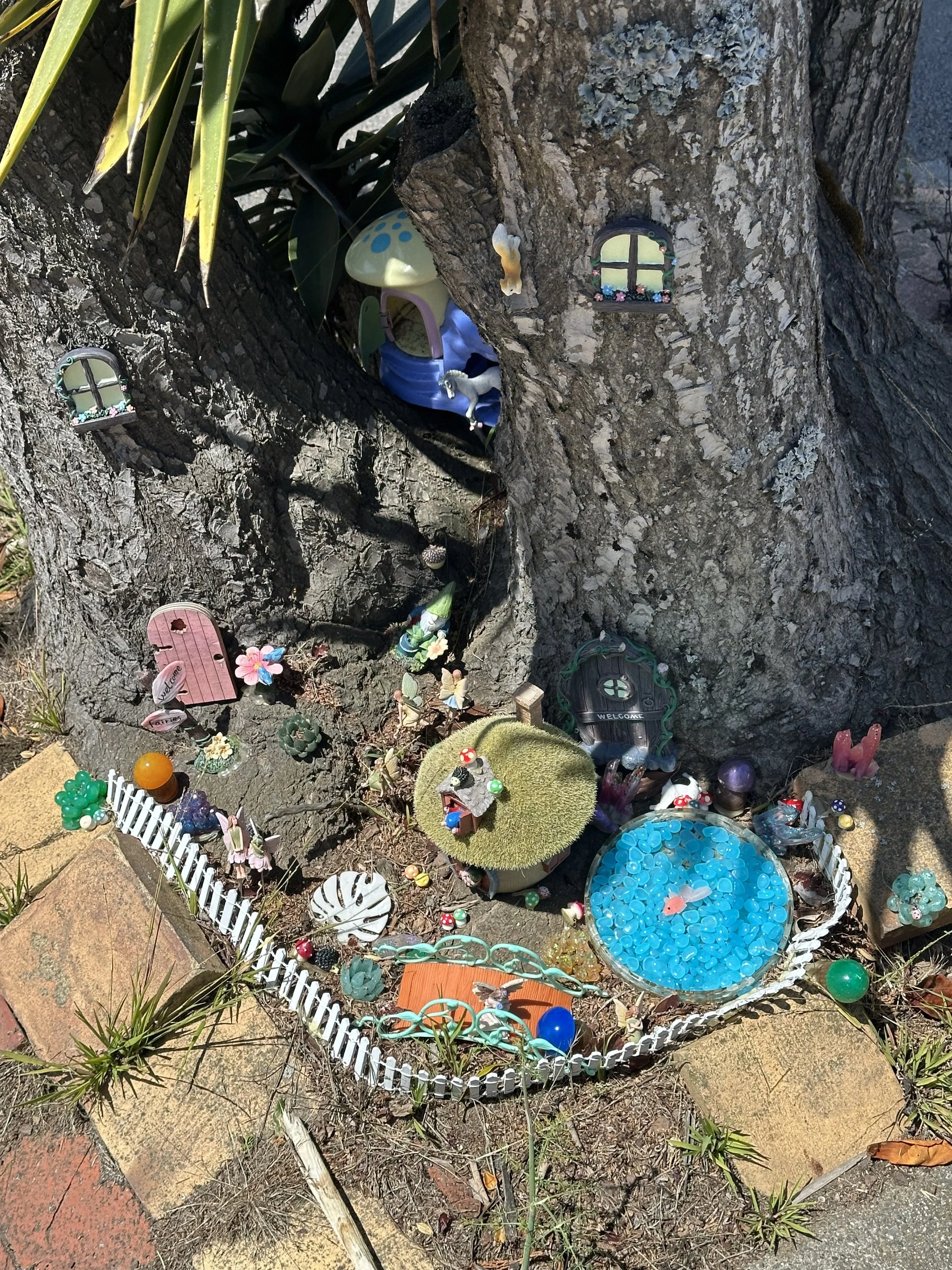 A backyard fairy garden with miniature fairy houses and decorations around a tree trunk, including small windows and a fairy door, with a small pond filled with blue stones and decorative ornaments.