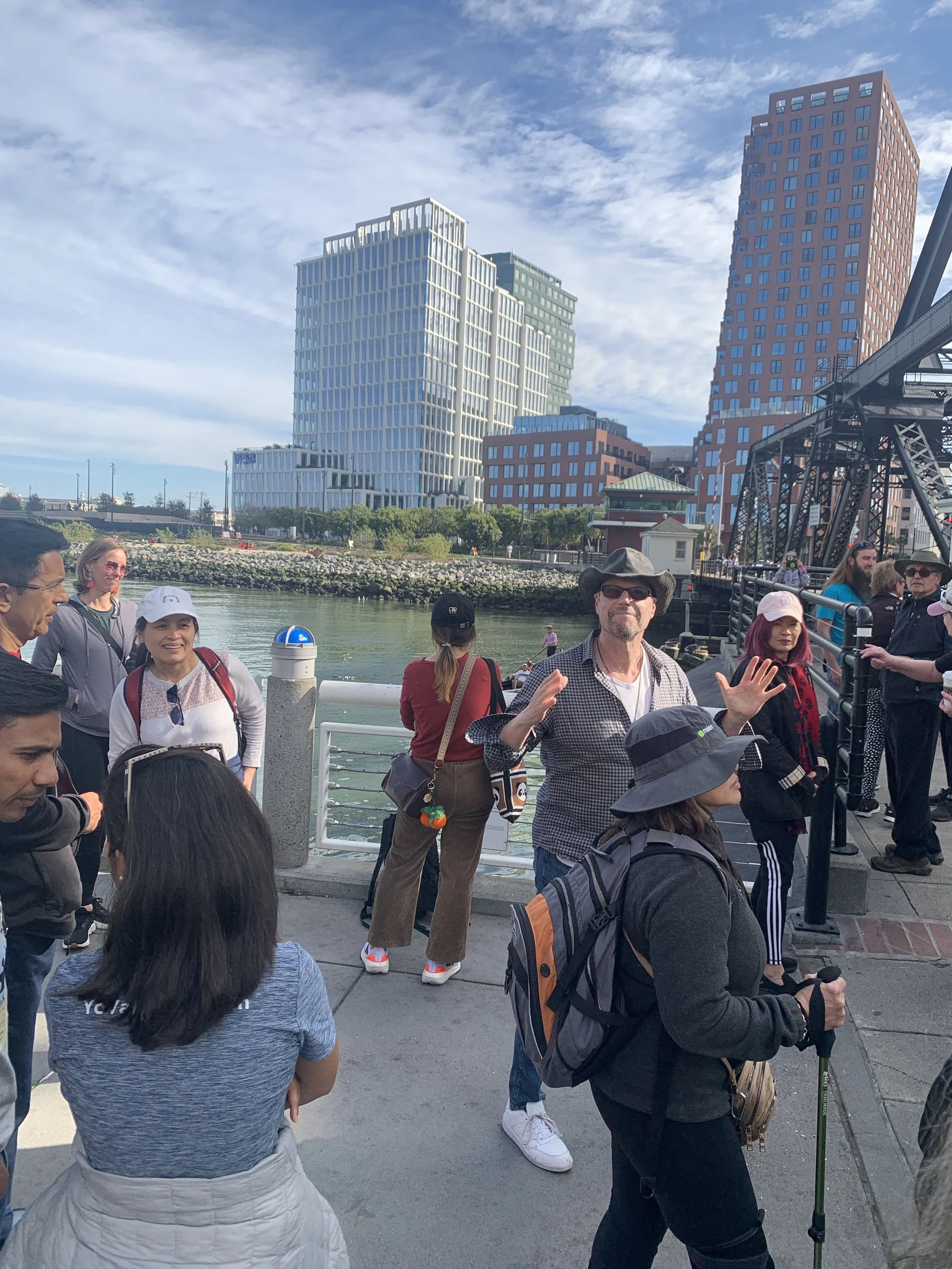 Group of people standing on a pier near the water with city buildings in the background, some smiling and talking, one person waving, some wearing hats and backpacks.