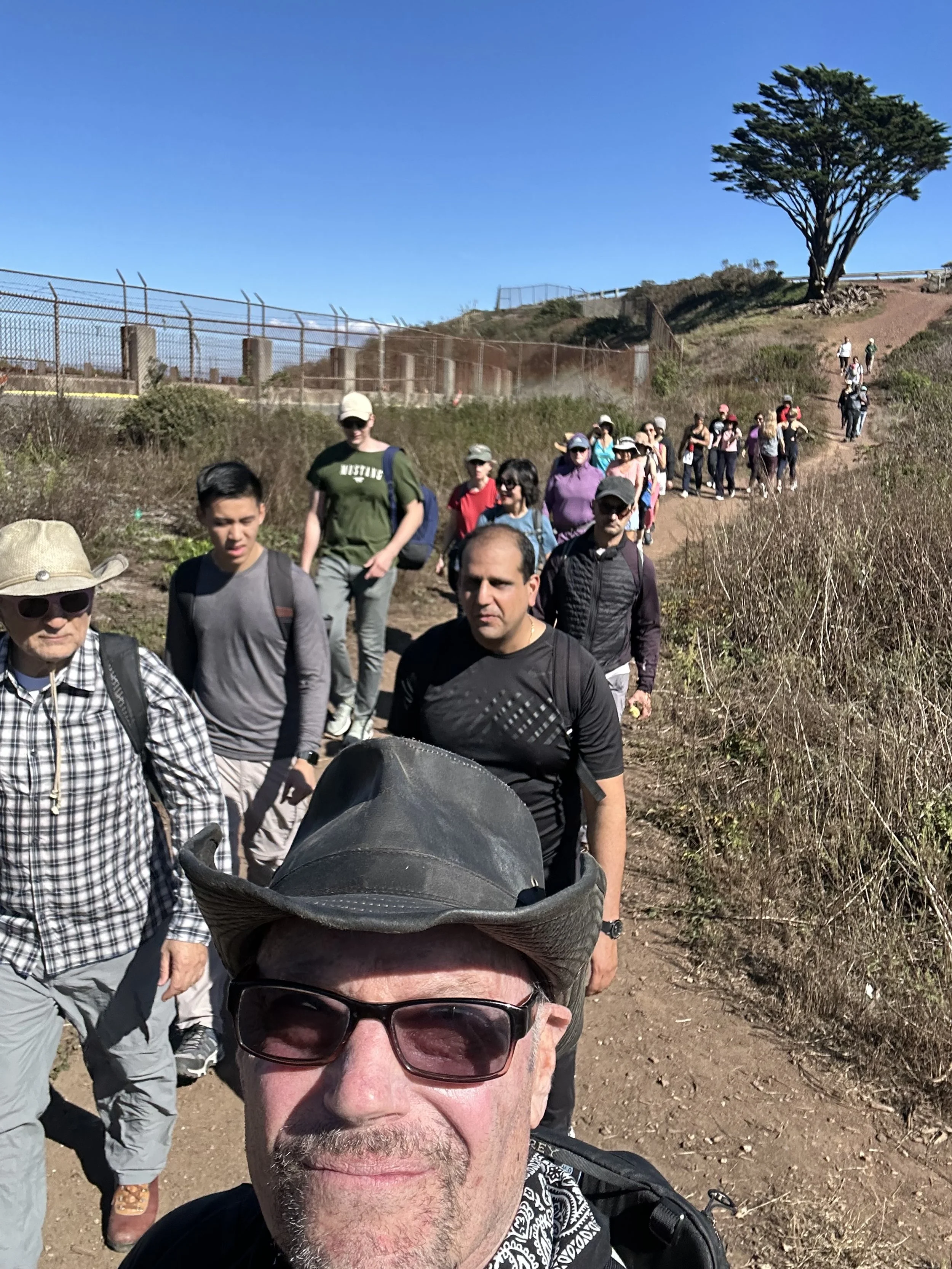 A diverse group of people hiking on a dirt trail in a hilly area with sparse vegetation and a large tree in the background on a clear, sunny day.