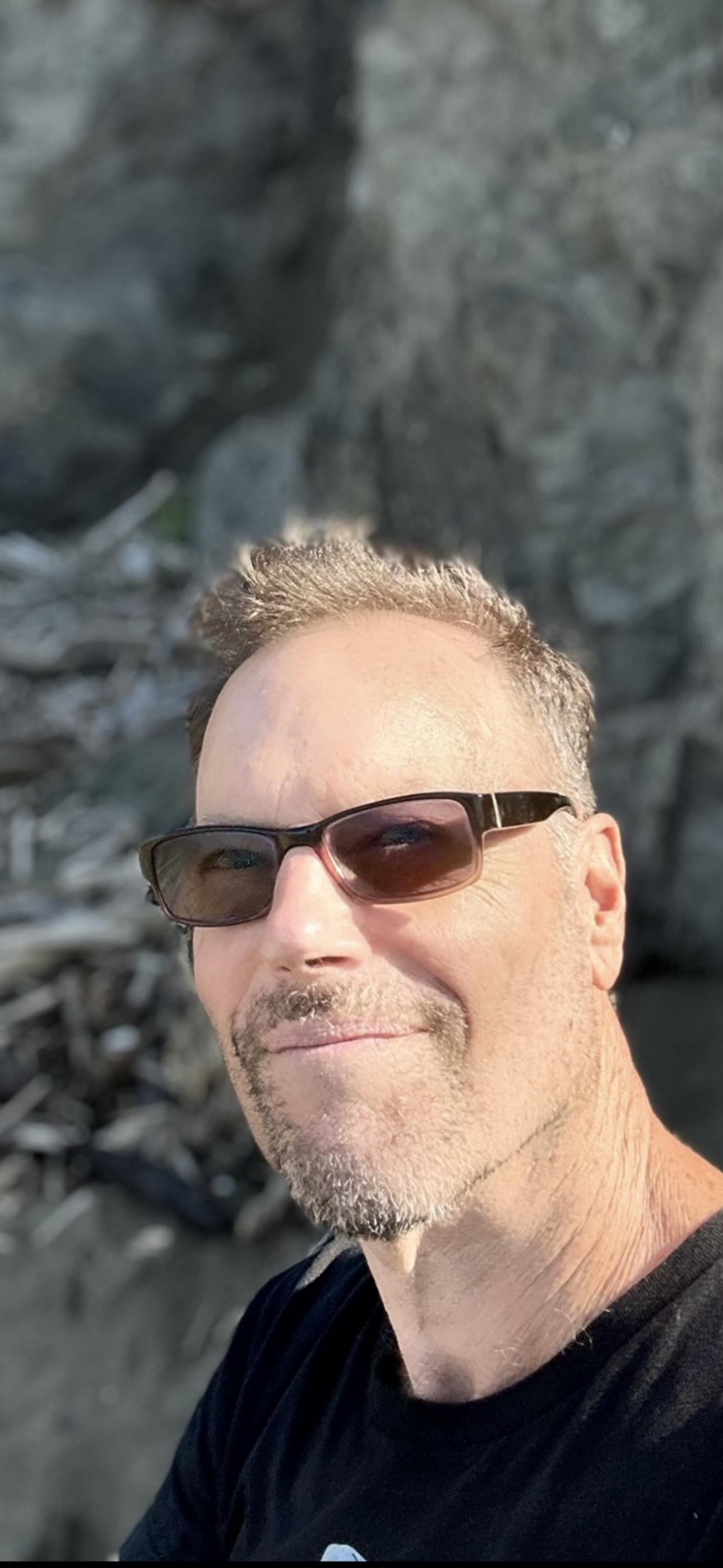 A man wearing sunglasses and a black shirt, smiling outdoors with a rocky background.