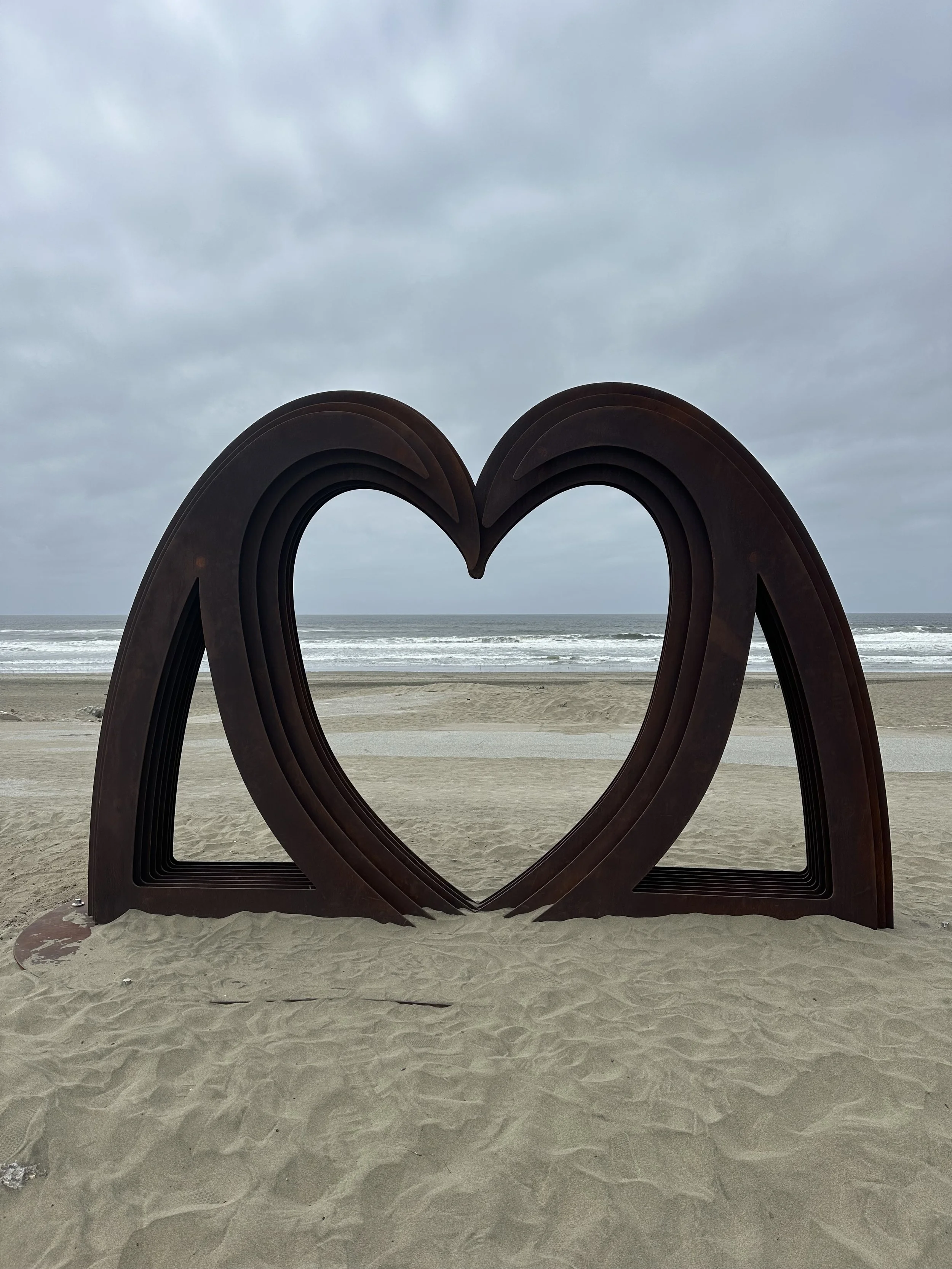 A large metal sculpture of two heart shapes leaning into each other on a sandy beach, with the ocean and cloudy sky in the background.