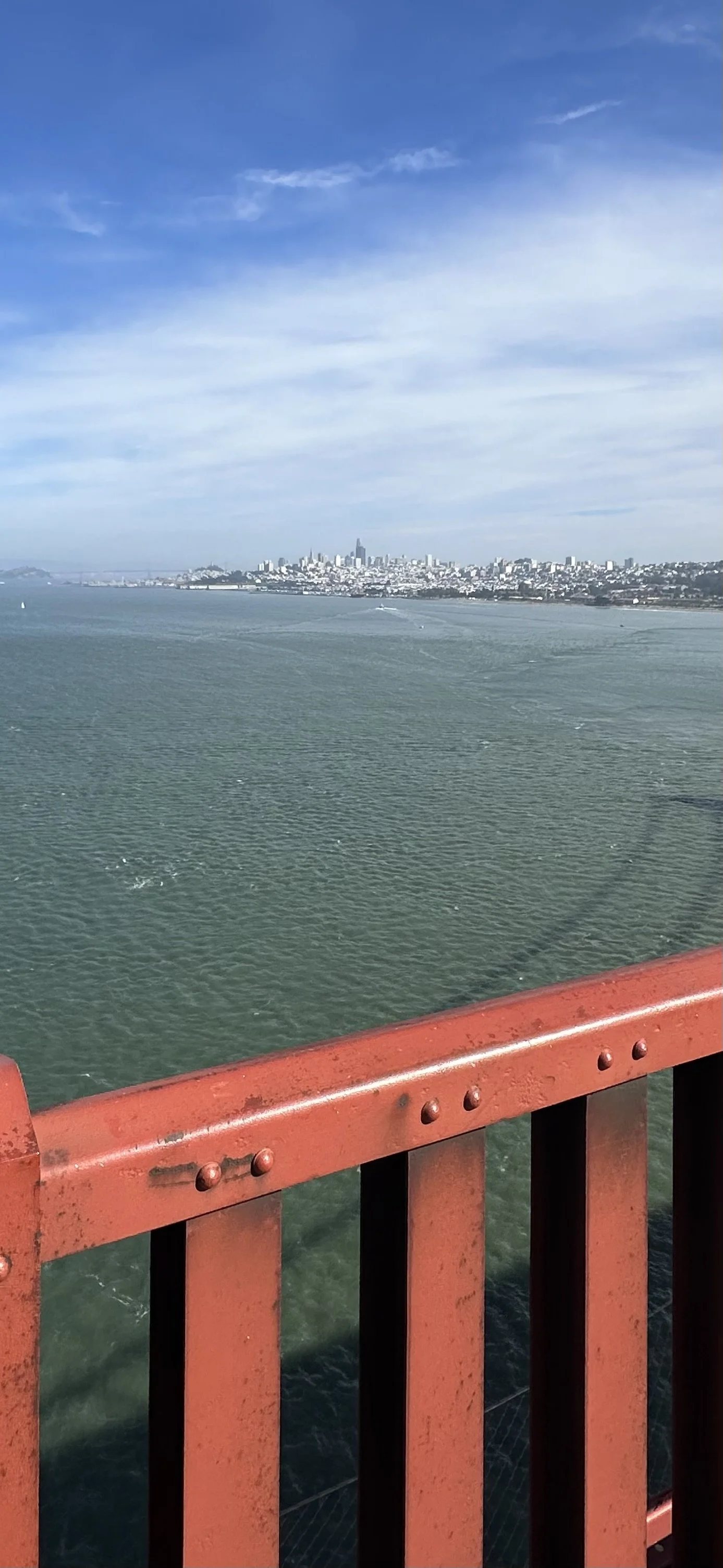 View of San Francisco skyline from a bridge, with water below and a red railing in the foreground.
