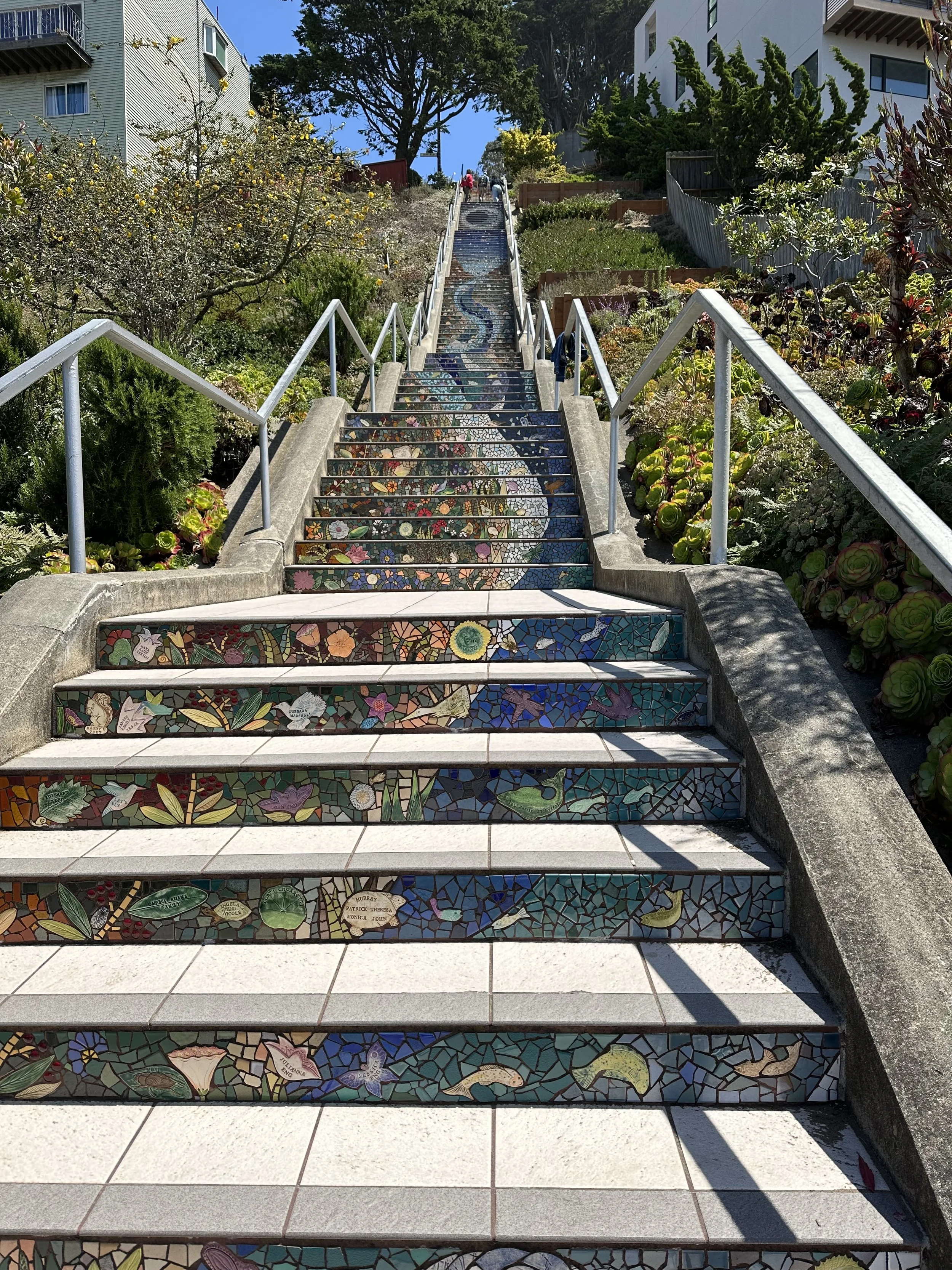 Colorful mosaic-covered stairs descending hillside, surrounded by lush plants and trees, with residential buildings in the background and a bright blue sky.