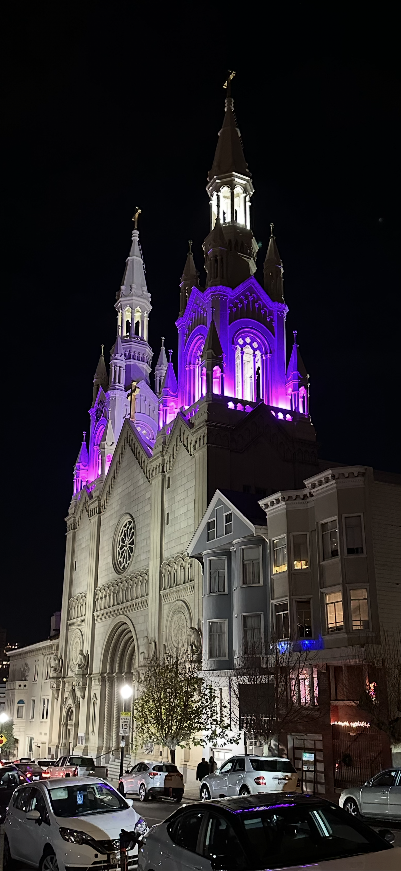 Night view of a tall, illuminated church with purple lighting accents, cars parked in front, and people walking nearby.