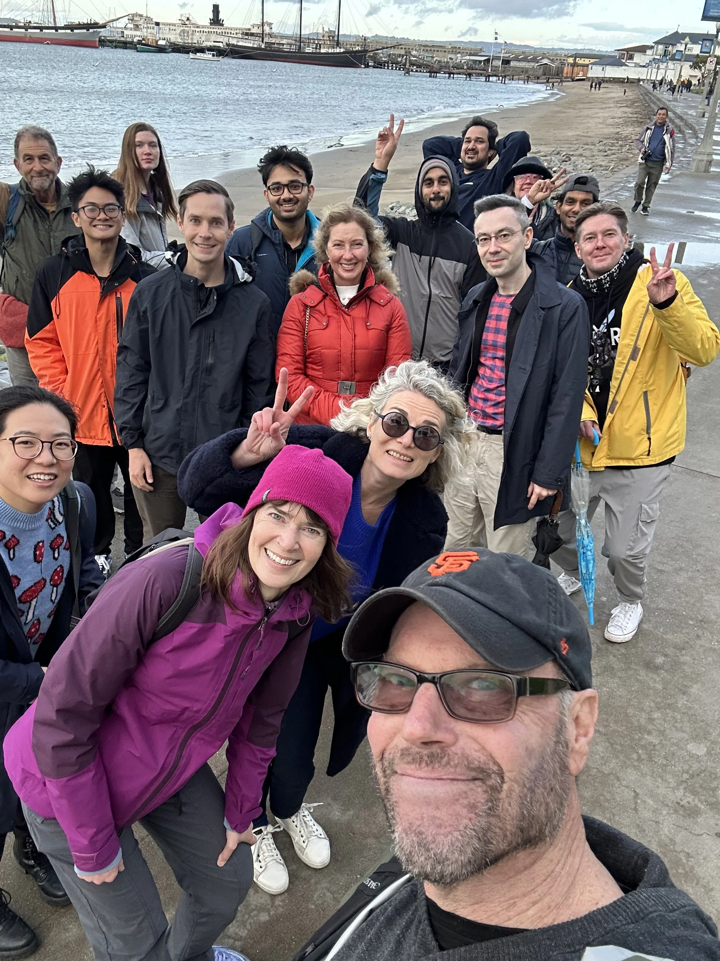 A group of smiling people taking a selfie on a waterfront promenade with ships in the harbor and buildings in the background.