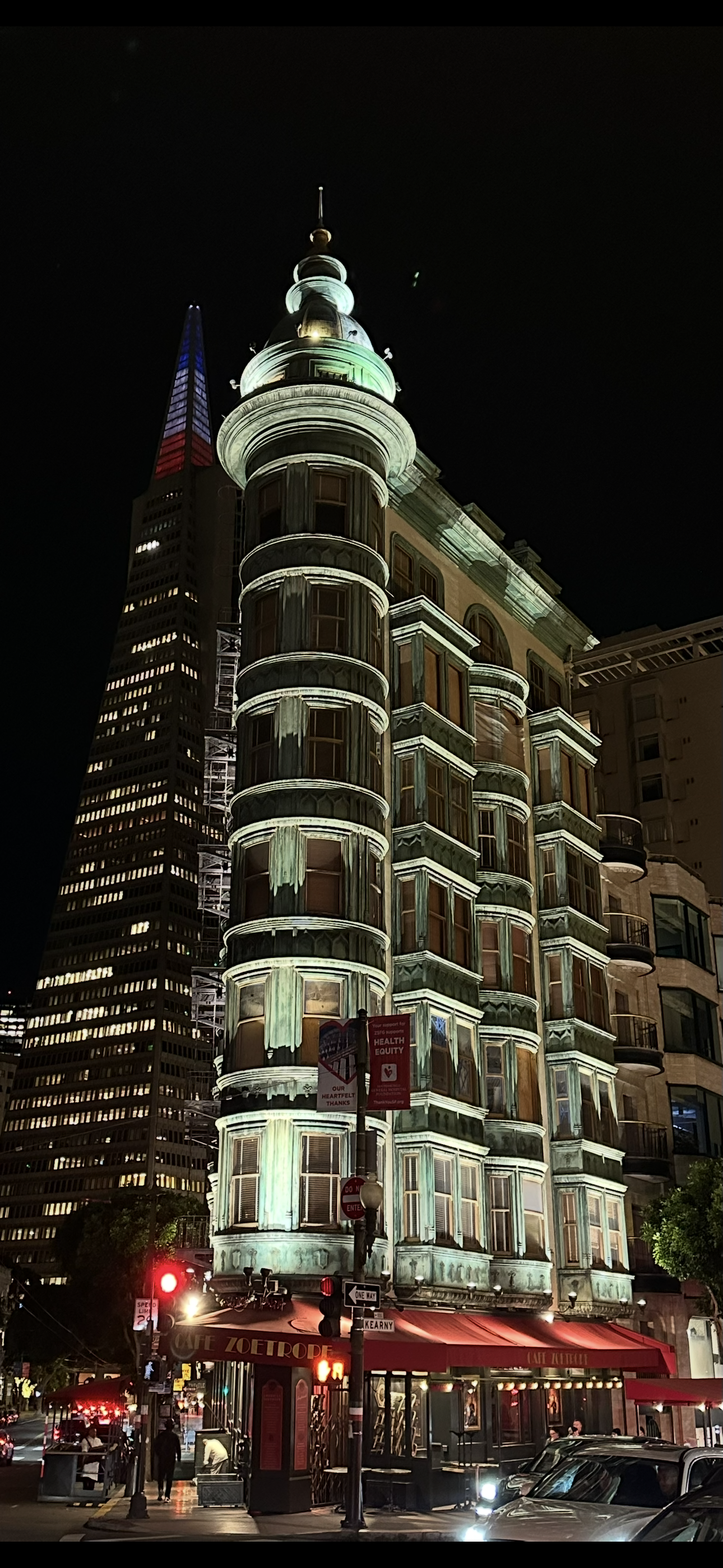 Night view of a tall, ornate building with a spire, illuminated at night, with a smaller skyscraper in the background, street traffic, and a red awning for Cafe Zoetrope at the corner.