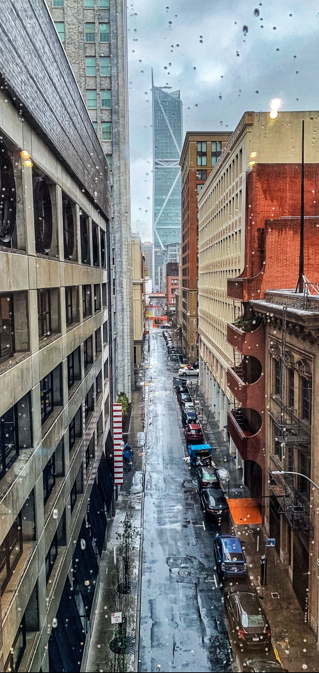 City street view from a rain-covered window, with parked cars lining the street, tall buildings on each side, and a modern skyscraper with diagonal structural elements in the background, under a cloudy sky.