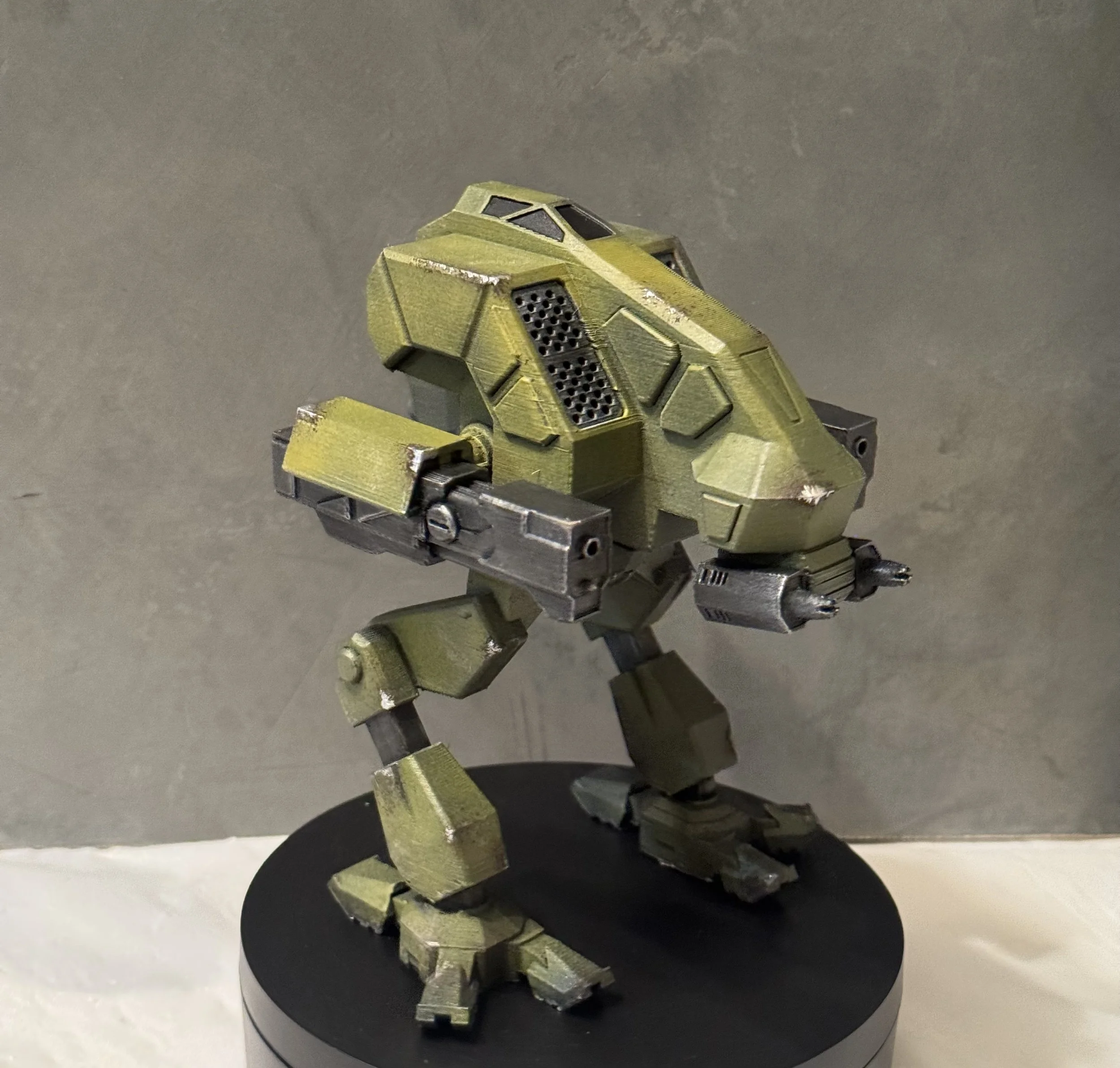 A detailed model of a futuristic military robot with a green camouflage pattern, standing on a round black stand against a gray background.