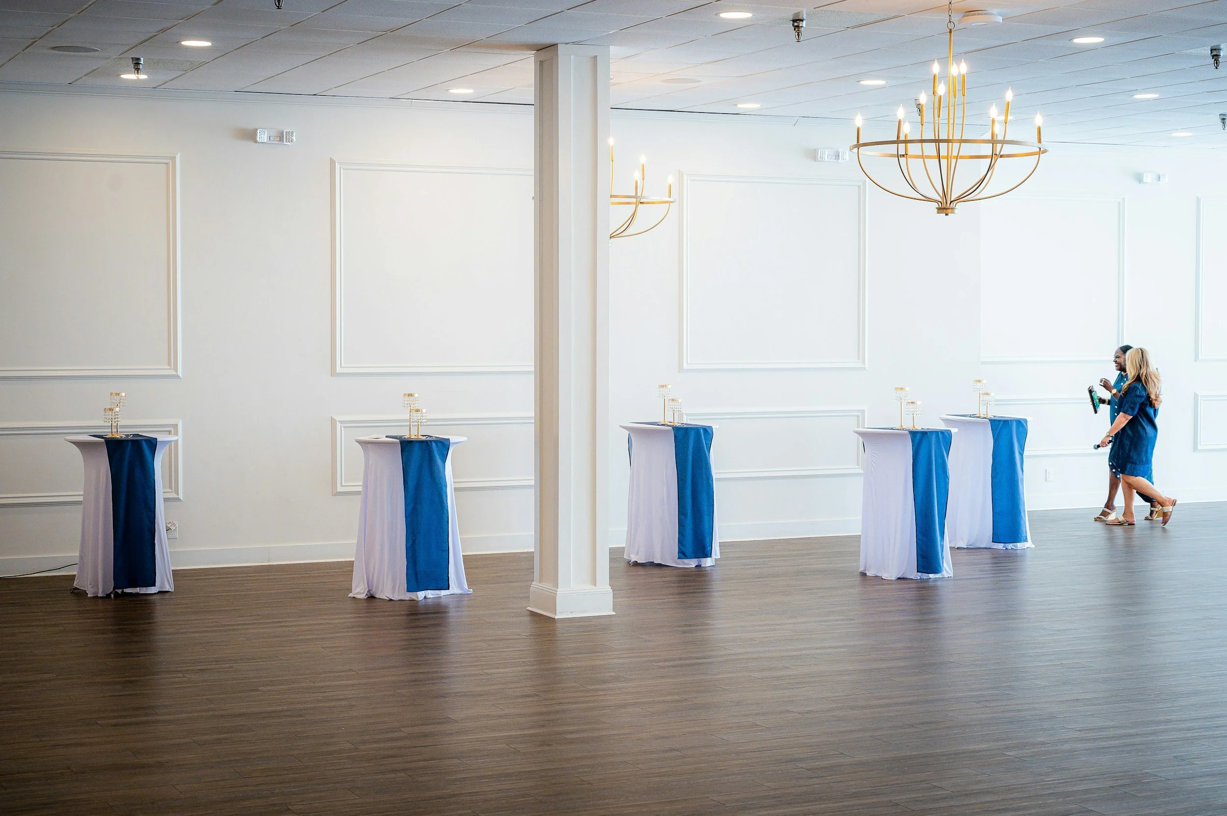 Empty event space with high-top tables covered in white and blue tablecloths, with two women walking in the background near a chandelier, in a room with white walls, wooden floor, and panel molding.