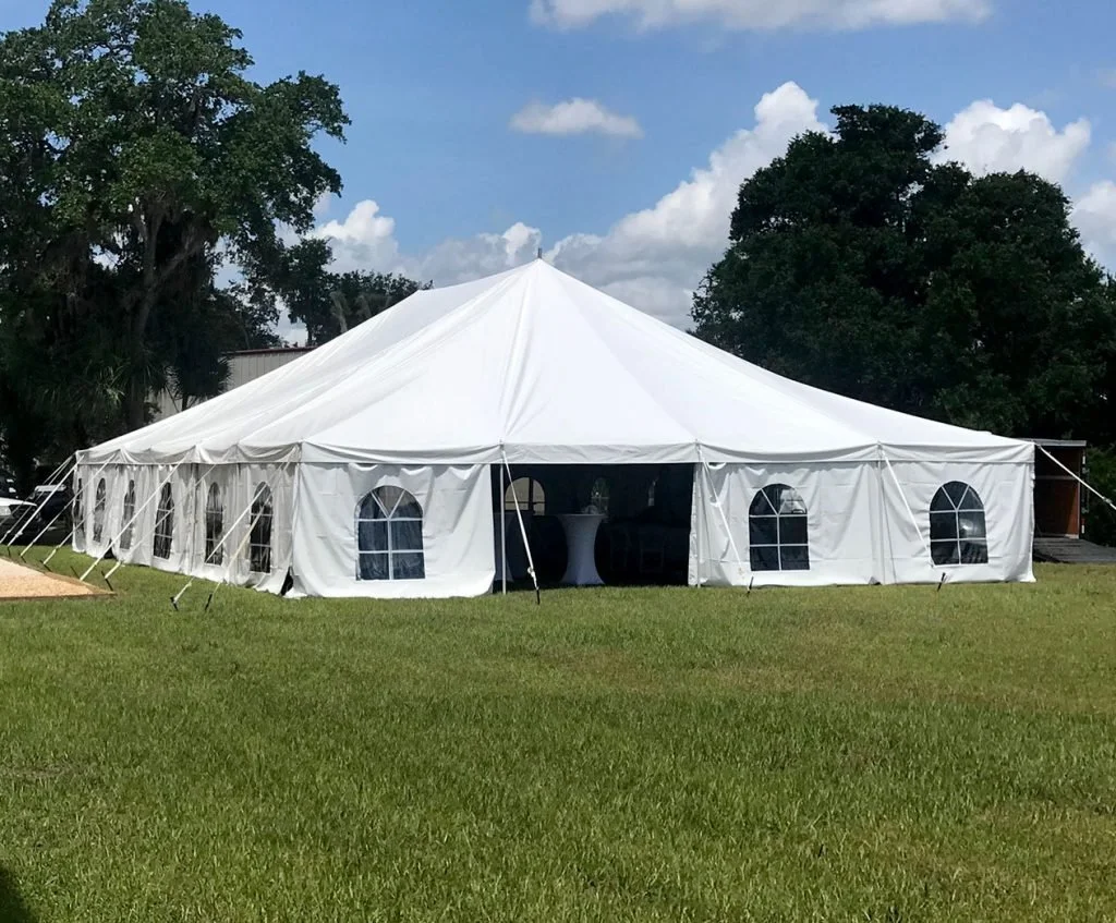 A large white event tent set up outdoors on a grassy area with trees in the background and a partly cloudy sky.