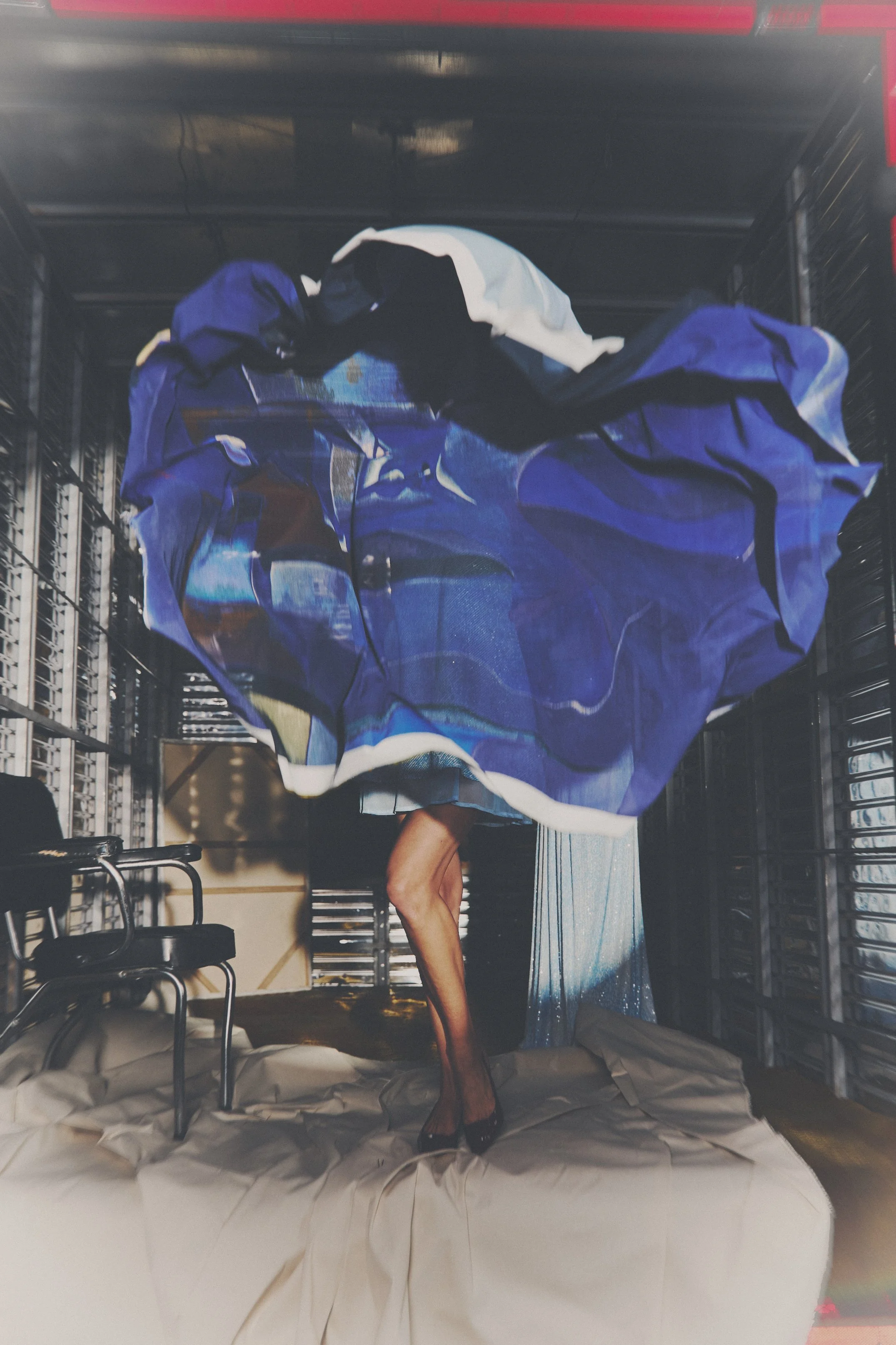 Una modelo con vestido azul y negro, con piernas desnudas y usando tacones negros, de pie sobre una cama arriba de sábanas blancas, en un espacio que parece un estudio o almacenamiento con paredes de metal y sillas negras.
