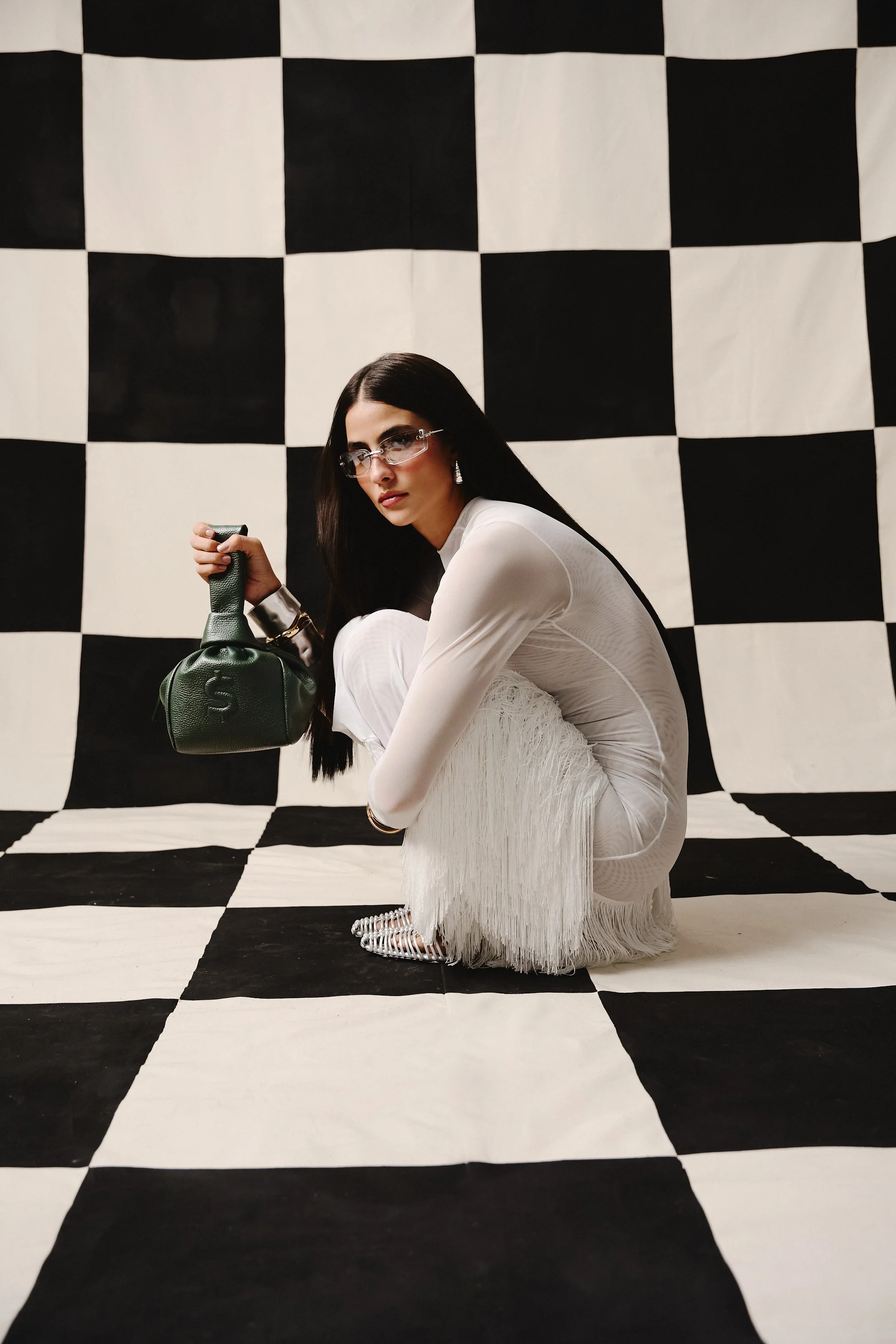 Mujer con cabello largo, vestido blanco con flecos, gafas y pendientes, agachada en un escenario con fondo y piso de patrón de tablero de ajedrez en blanco y negro, sosteniendo un bolso verde.